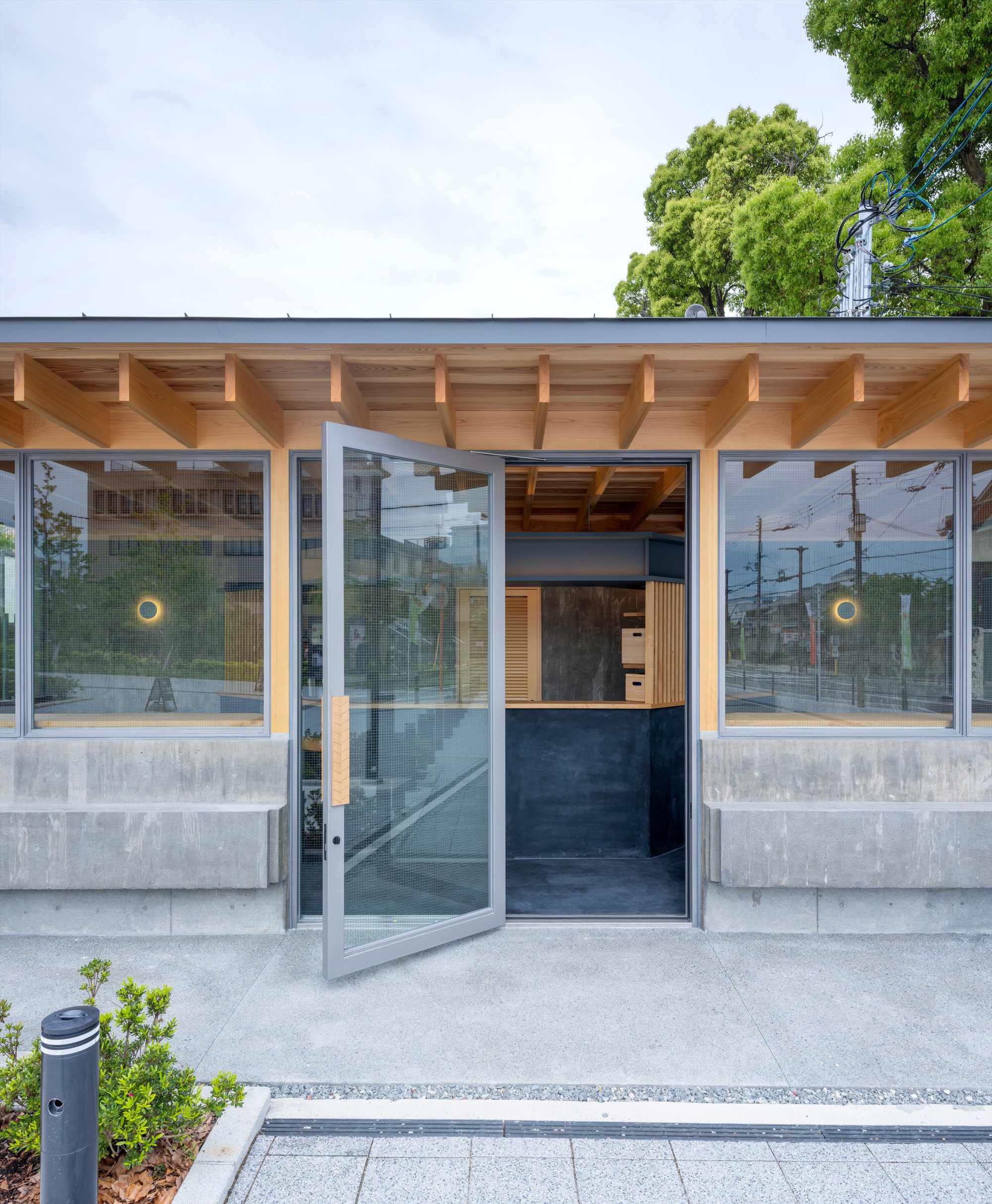 Open pivot glass door in the kiosk front, with exposed timber rafters and concrete benches under a cloudy sky