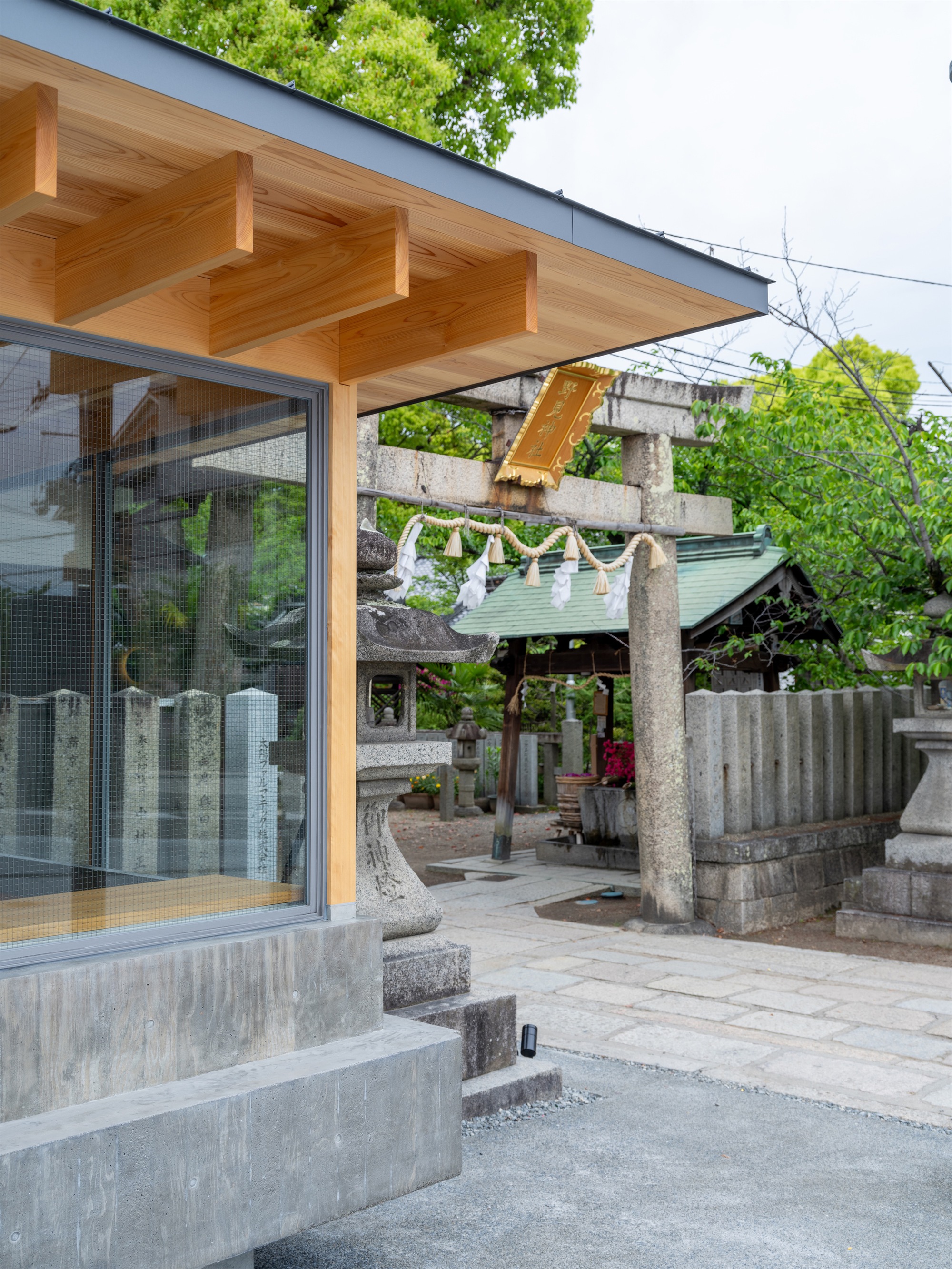 Corner of the kiosk with exposed timber rafters beside a stone torii gate hung with a braided straw rope, in green foliage
