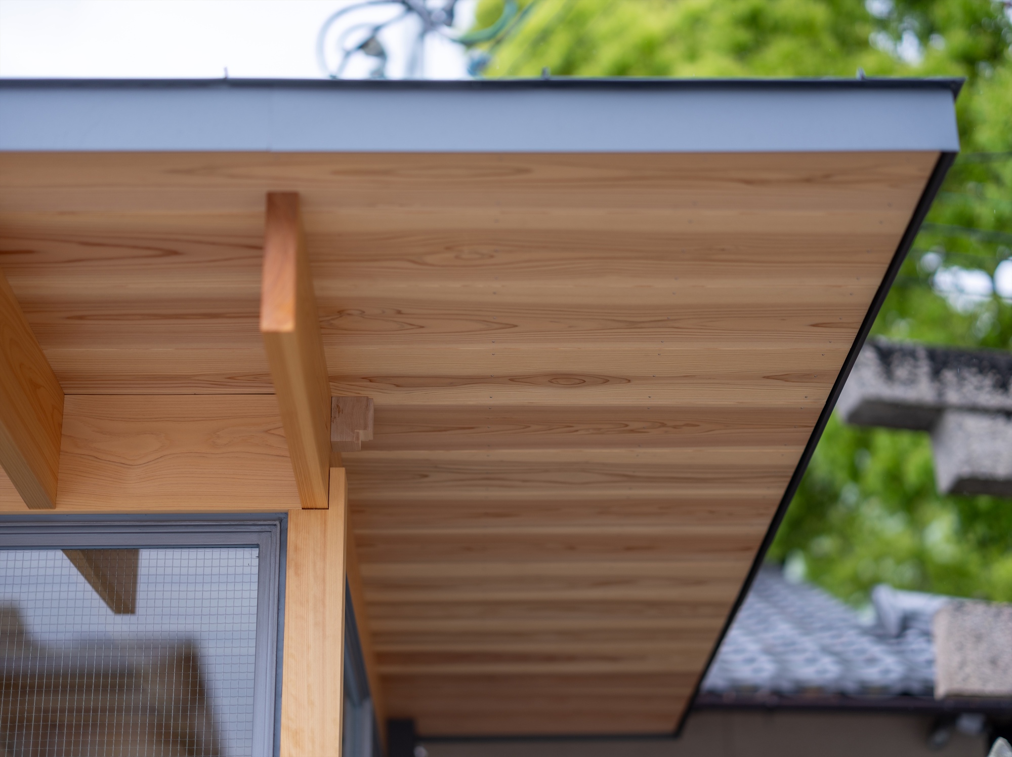 Underside of the cedar-boarded eave with a projecting rafter end and grey metal fascia, framed by green foliage and a tiled roof beyond