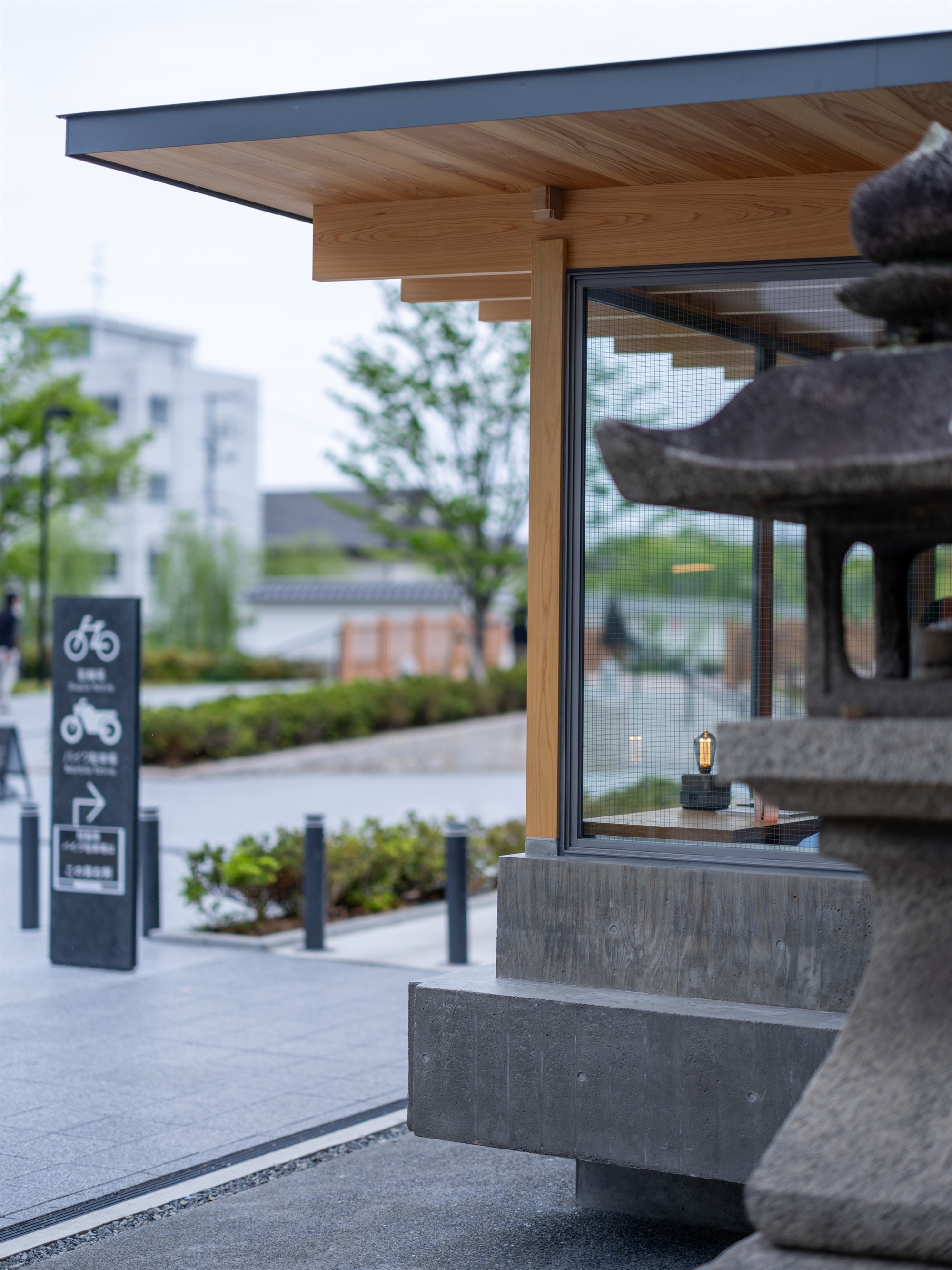 Glazed corner of the timber pavilion beside a stone lantern and a bicycle sign, with a concrete base under an overcast sky
