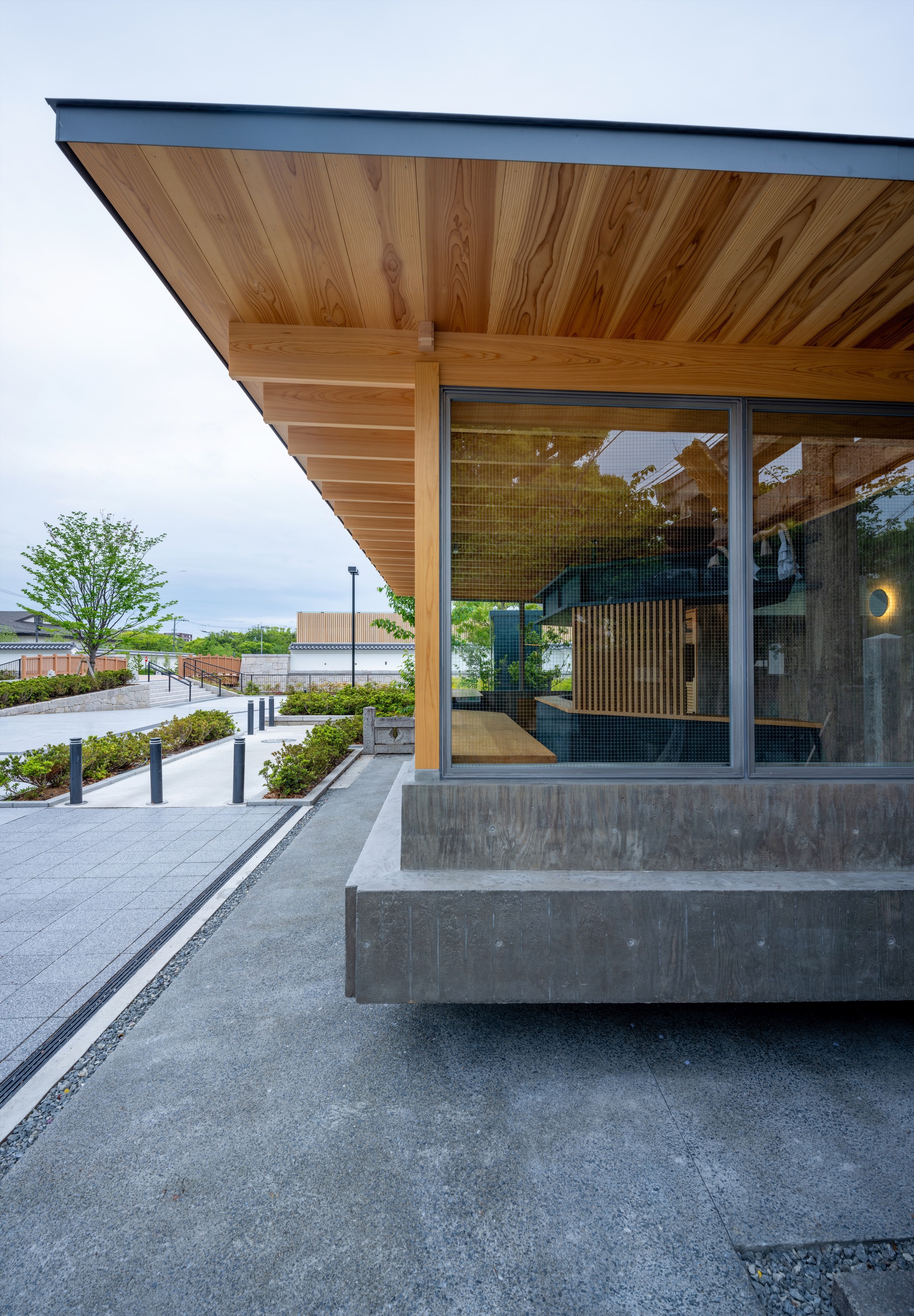 Corner of the pavilion with cantilevered cedar eave, board-formed concrete plinth and wire-mesh glazing above a paved path at dusk