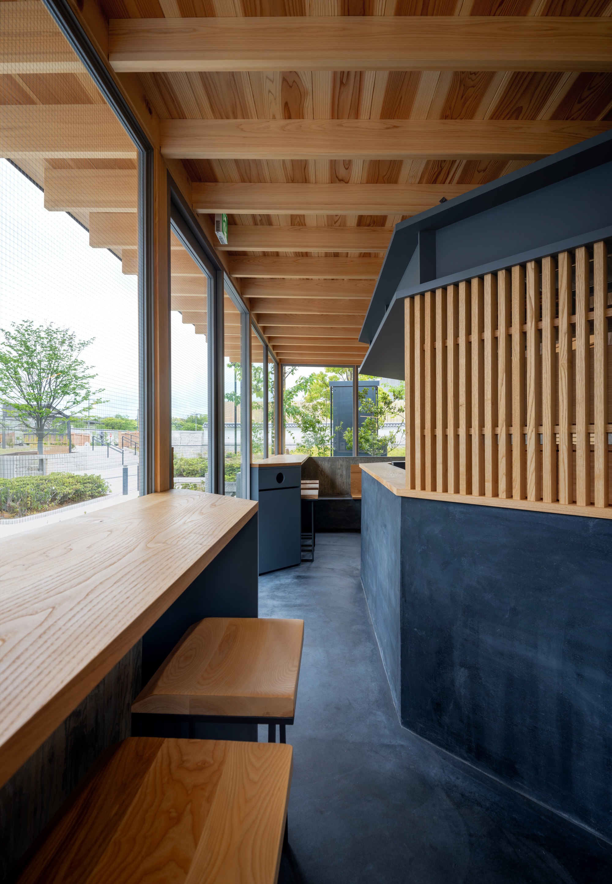Interior view along a window counter with timber stools, a slatted wood screen and a dark plastered service counter beneath exposed beams