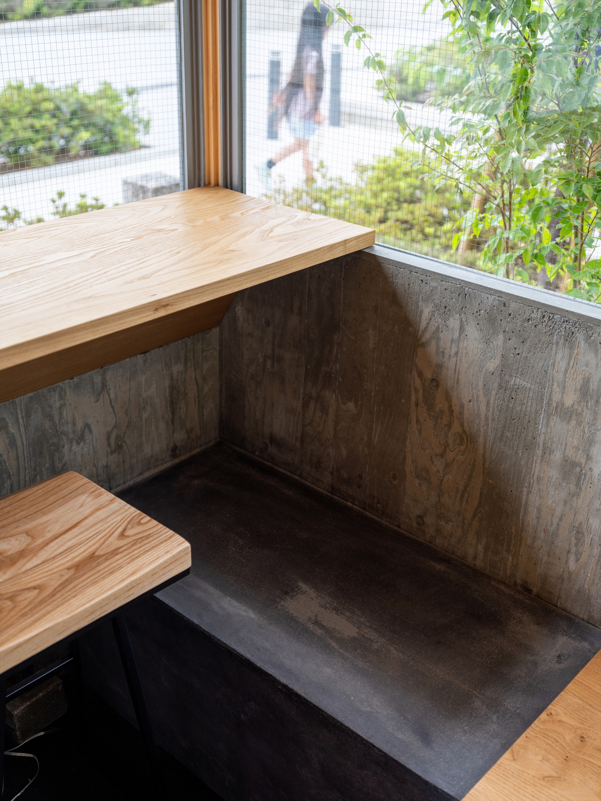 Window corner with an oak counter and a concrete bench, while a person walks past outside the wire-mesh glass