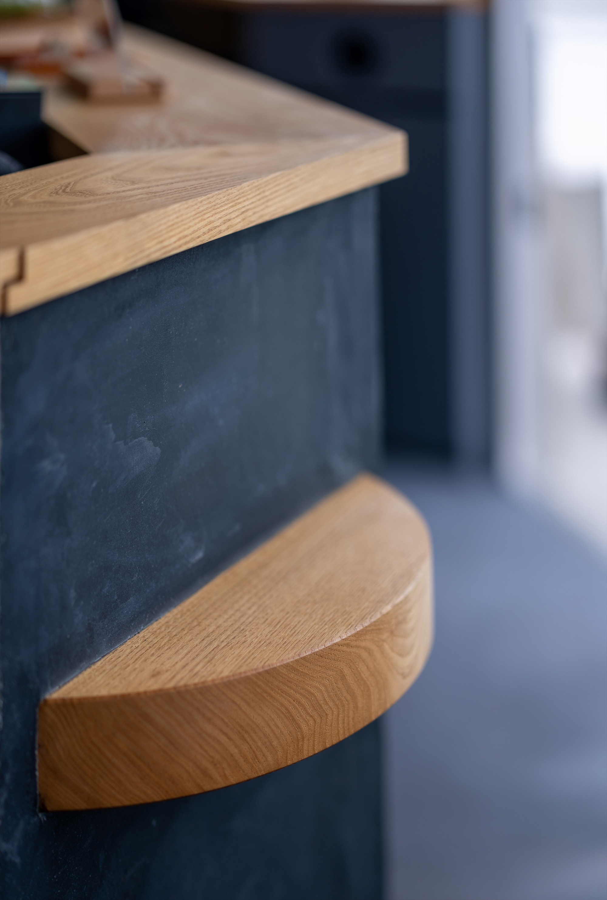 Close-up of a half-round oak step projecting from a dark slate-clad counter, with a blurred grey floor behind