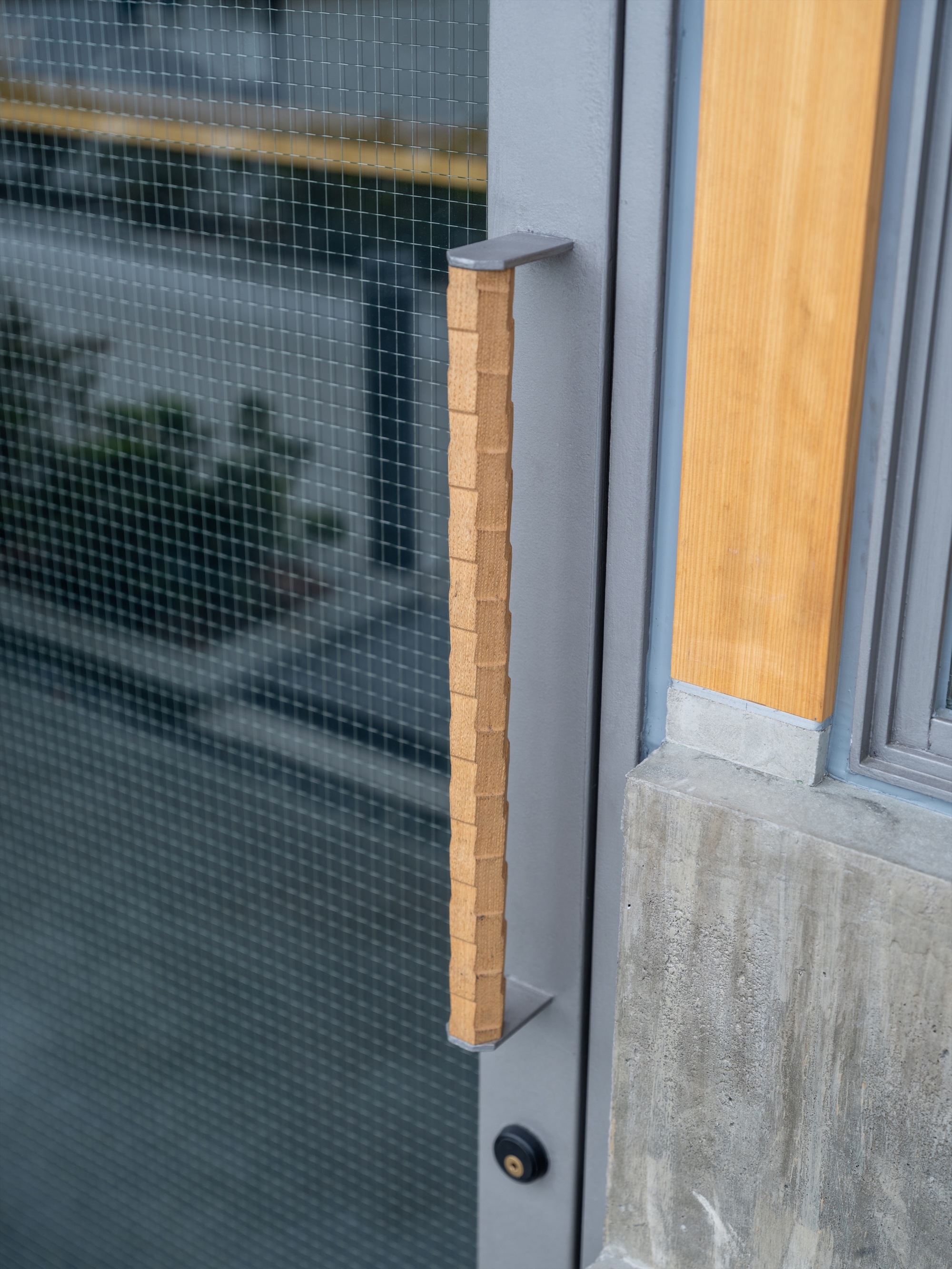 Vertical oak handle with notched surface on a grey steel glass door, board-formed concrete base beside it