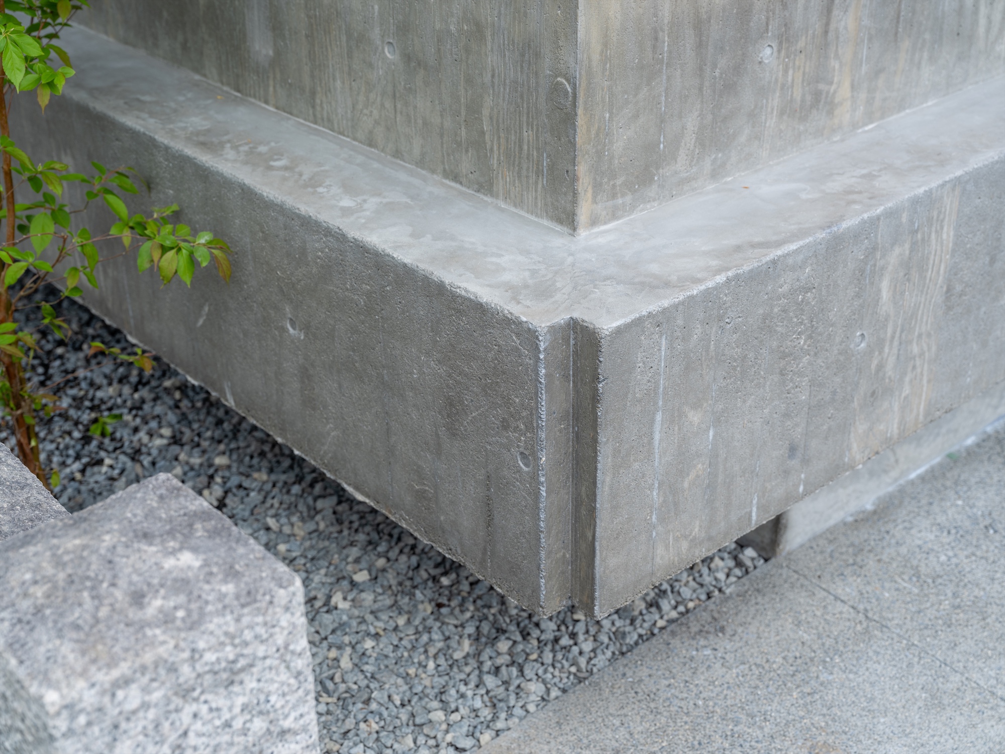 Corner of a board-formed concrete plinth over grey gravel, with a young shrub and granite block at the edge