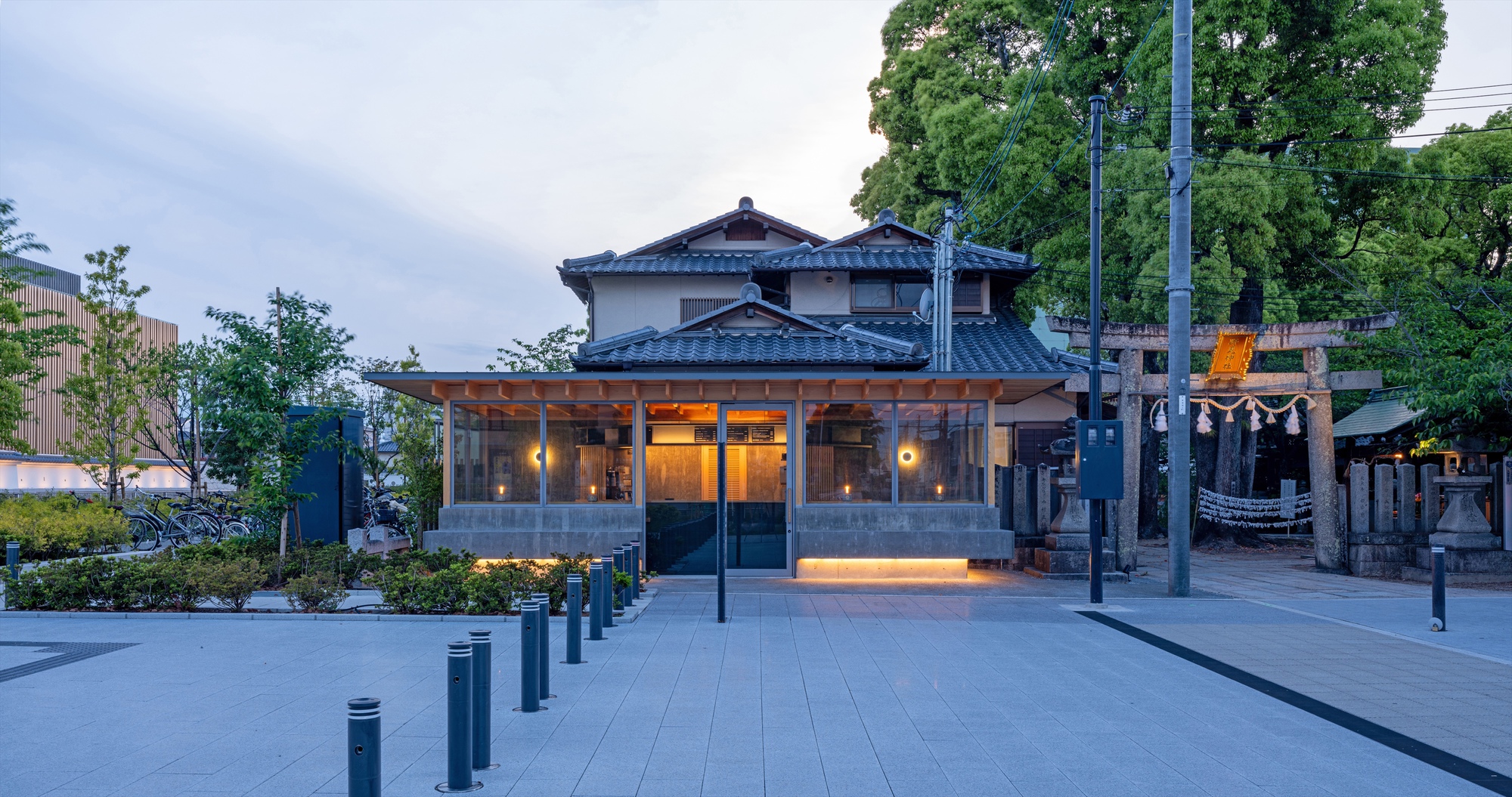 Glazed timber-framed pavilion with glowing base lighting in front of a tiled-roof house and shrine gate at dusk