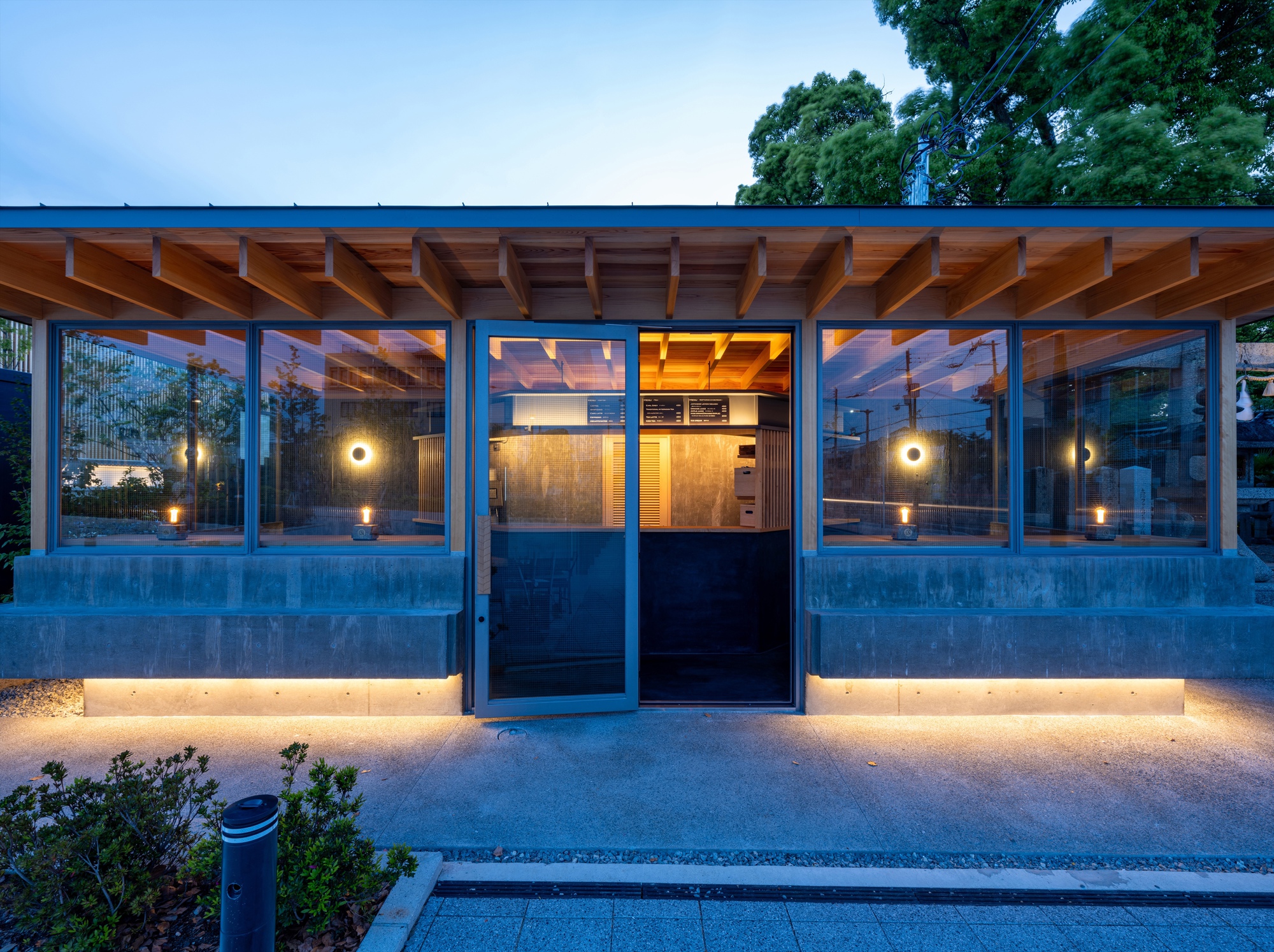 Front of the glazed pavilion with exposed timber rafters, open door and lit concrete base at blue hour
