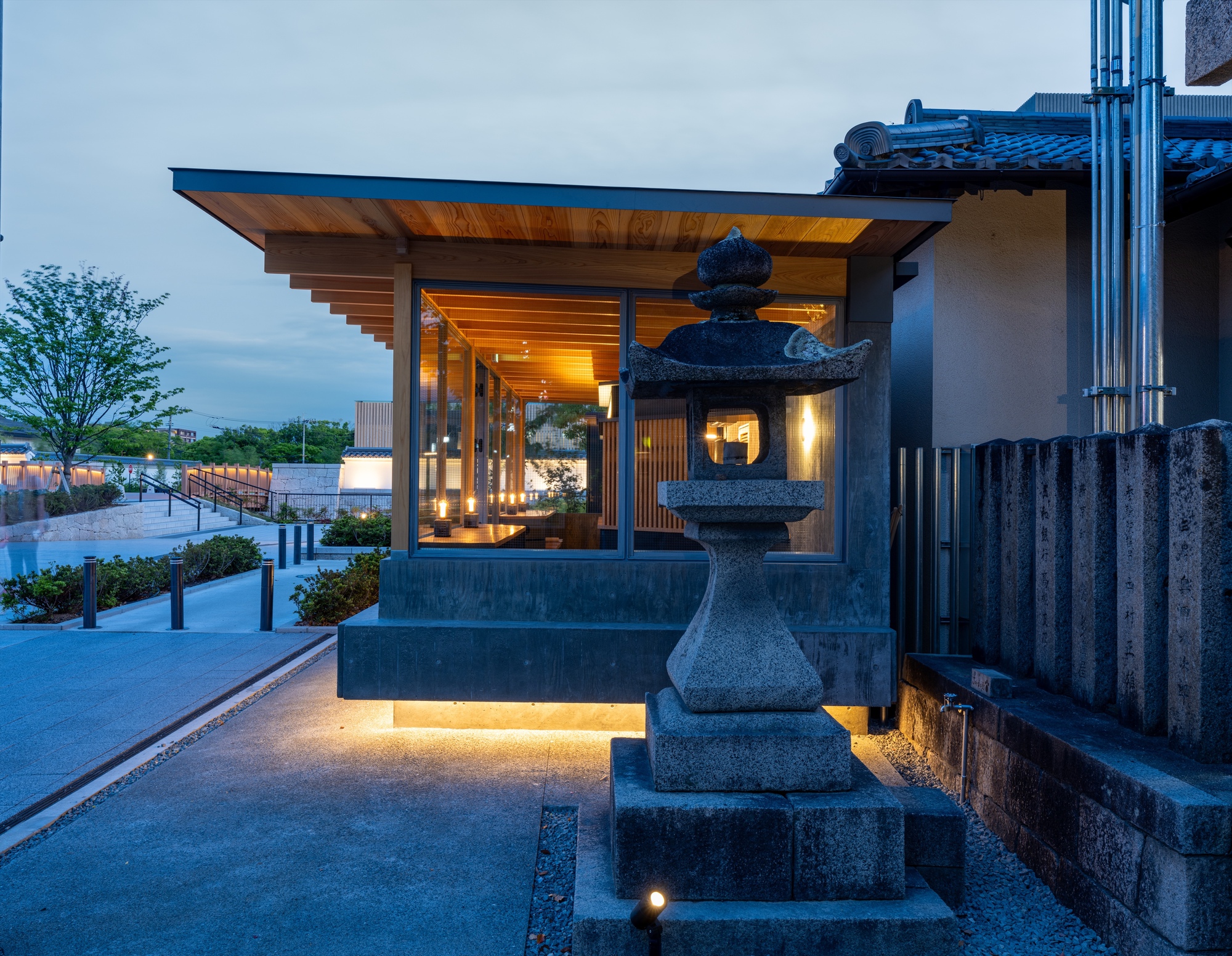 Small glazed pavilion with a timber soffit and stone lantern in the foreground, lit by warm lights at dusk