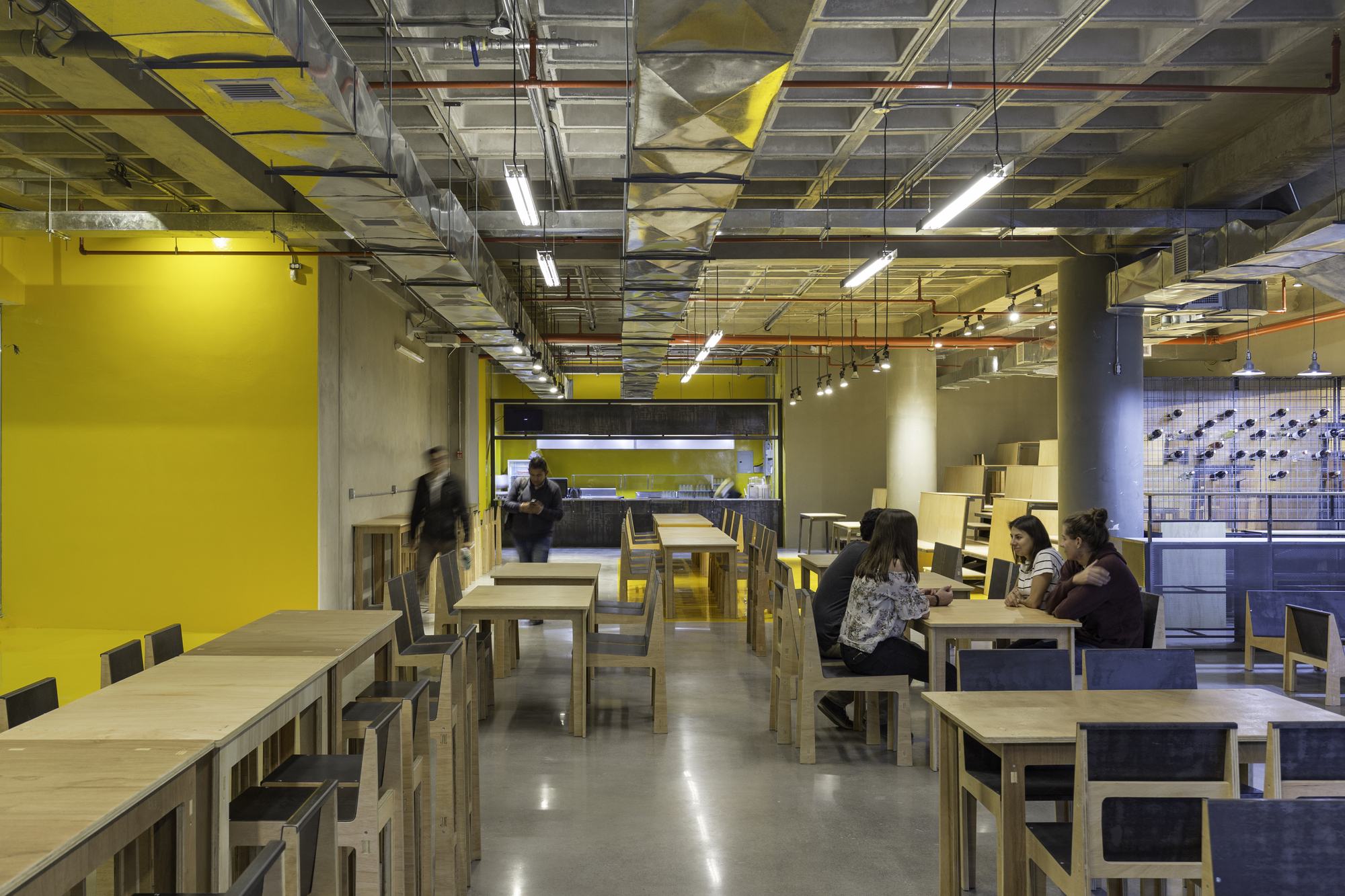 Open dining hall with plywood tables and chairs, a yellow wall and exposed ducts, with diners seated and one person walking
