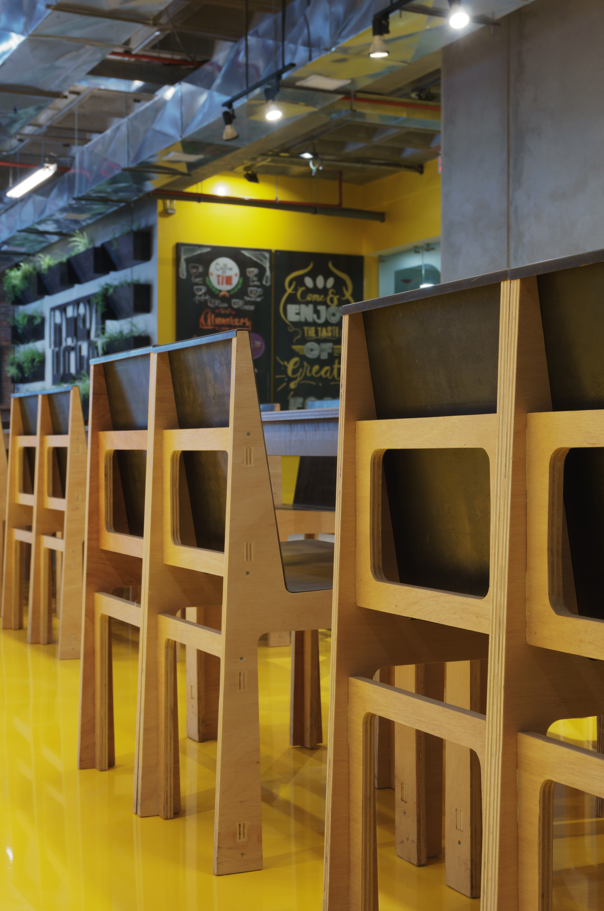 Close-up of plywood chair backs with cutout openings on a glossy yellow floor, chalkboard menus glowing behind