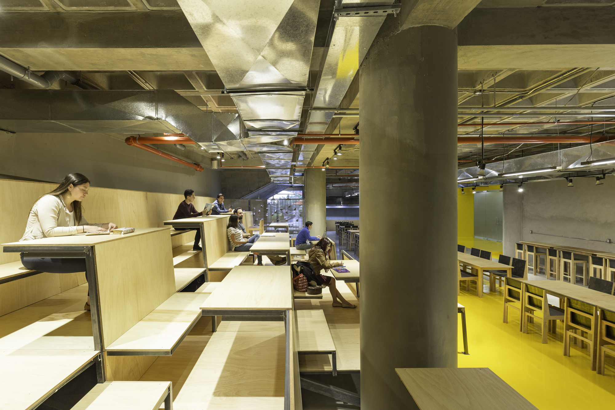 Stepped timber seating platform where people read and work at long tables beside a concrete column, with exposed ducts and a yellow floor beyond