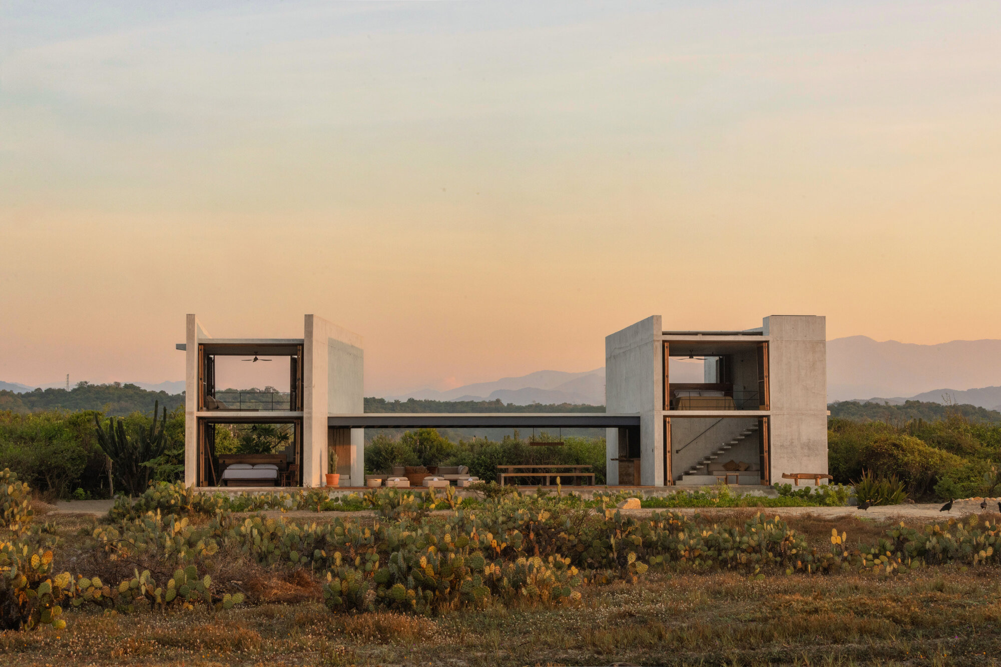 Two concrete pavilions joined by a low bridge beam, with open bedrooms and a stair, set among prickly pear at sunset