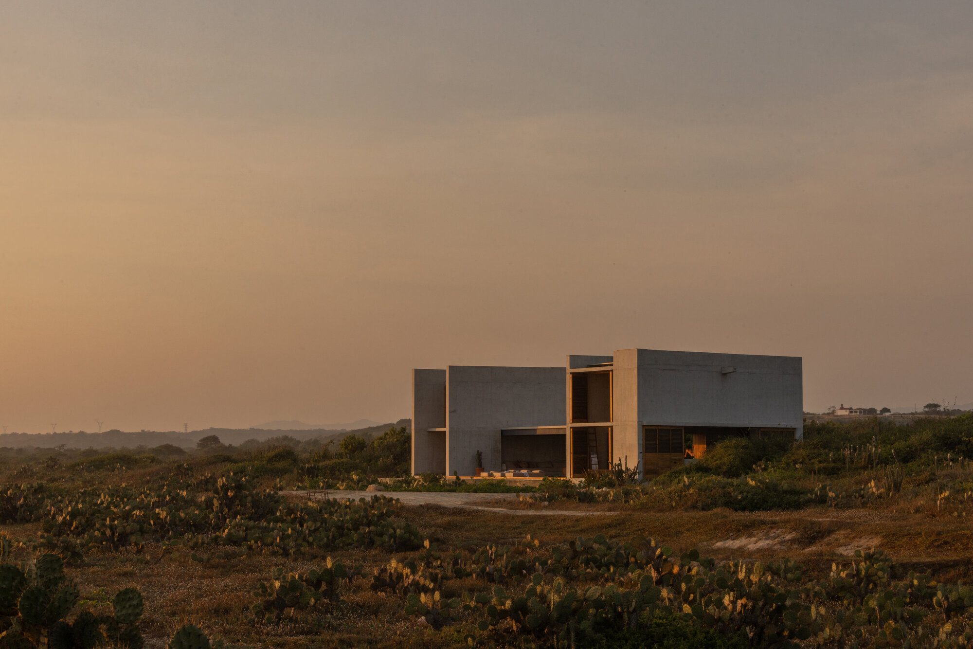 Distant view of the blocky concrete house with open corners, surrounded by cactus scrub under a hazy orange dusk sky