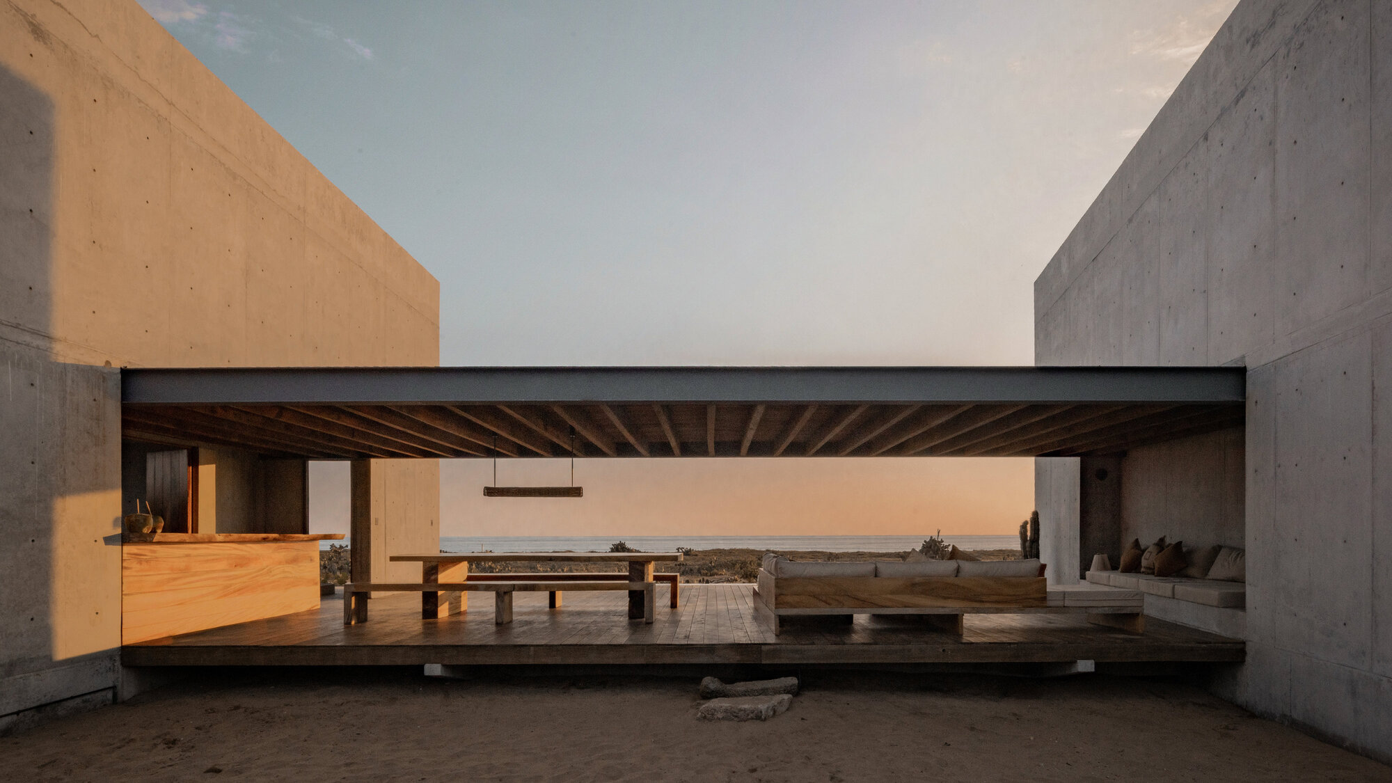 Open-sided living pavilion between two concrete walls, with a timber deck, long dining table and built-in sofa facing the sea at dusk