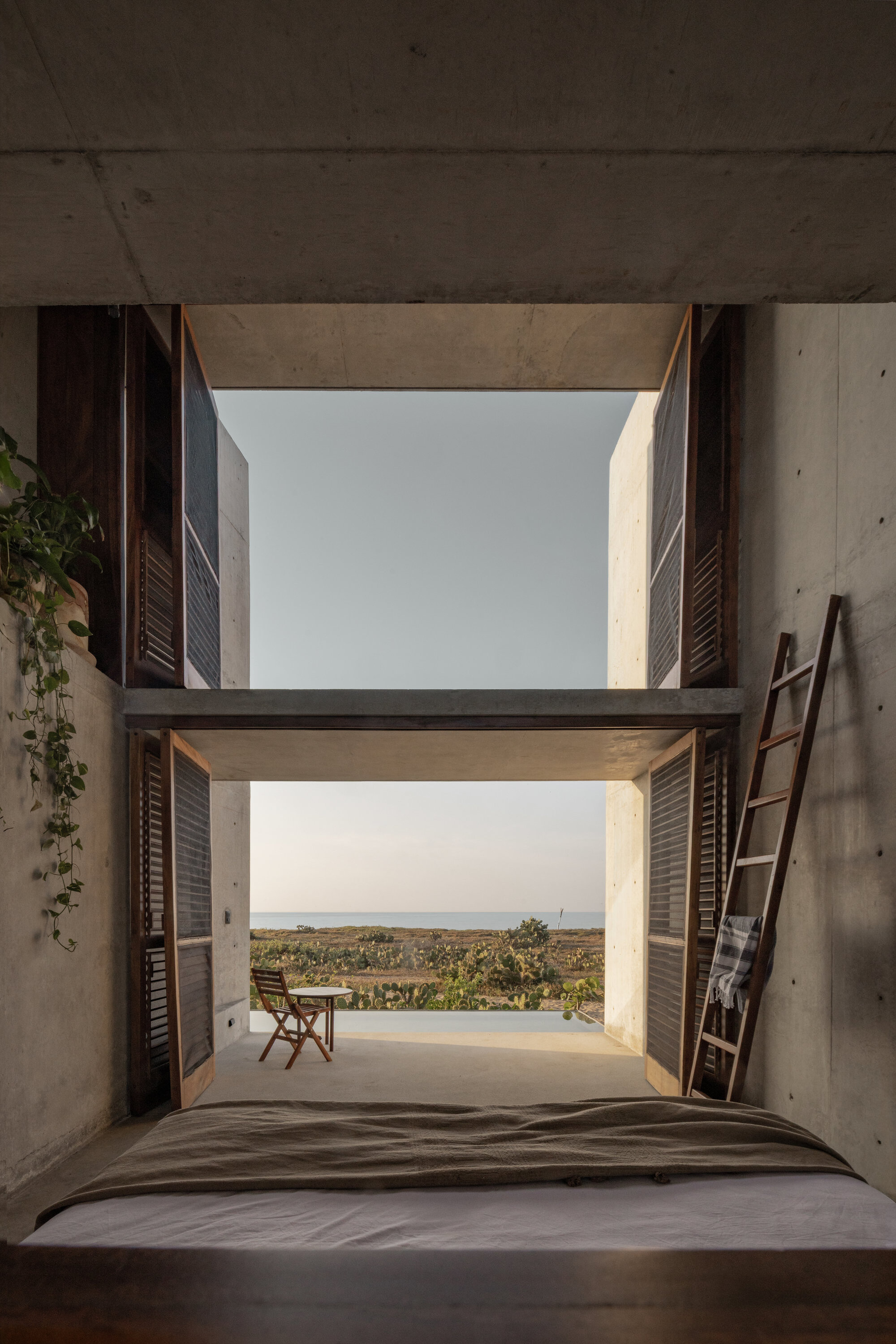 Bedroom opening through folded wooden shutters to a terrace chair and the horizon, with a leaning ladder and trailing vine