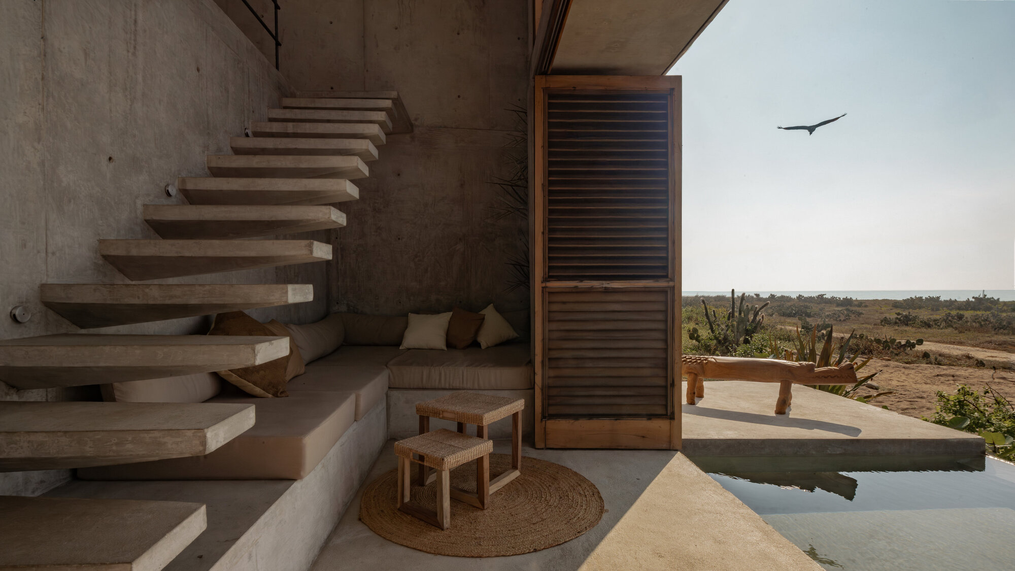 Cantilevered concrete stair above a built-in seat and woven rug, with a bird flying over the plunge pool and scrub beyond