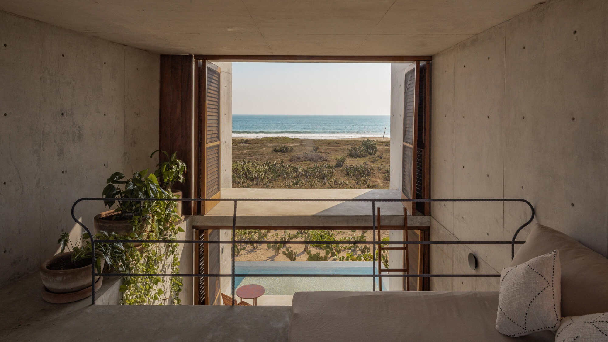 Concrete mezzanine with a daybed and potted plants, framed by open timber shutters facing a pool and the distant ocean