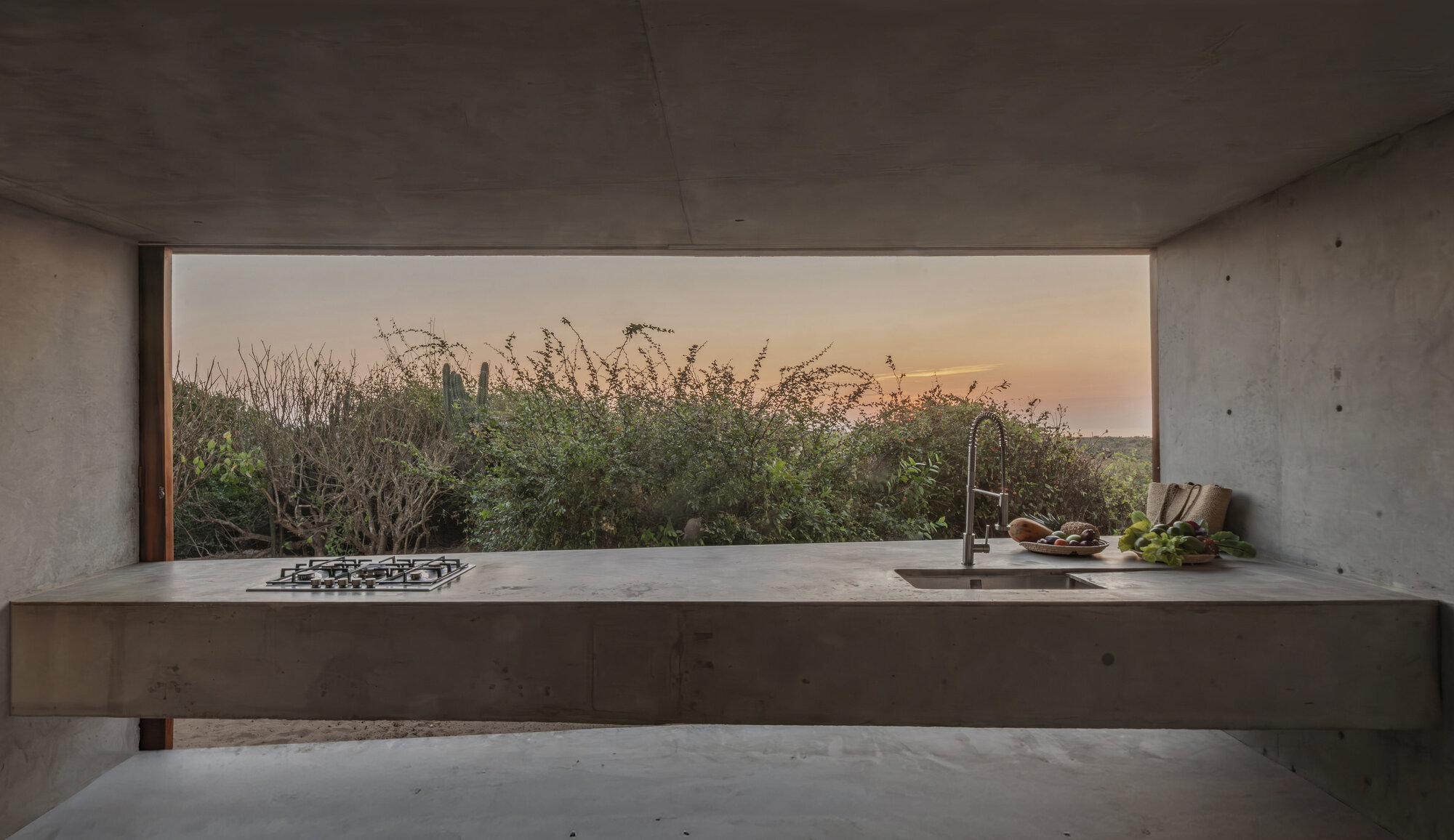 Cantilevered concrete kitchen counter with a gas hob and sink, framed against scrub vegetation at sunset