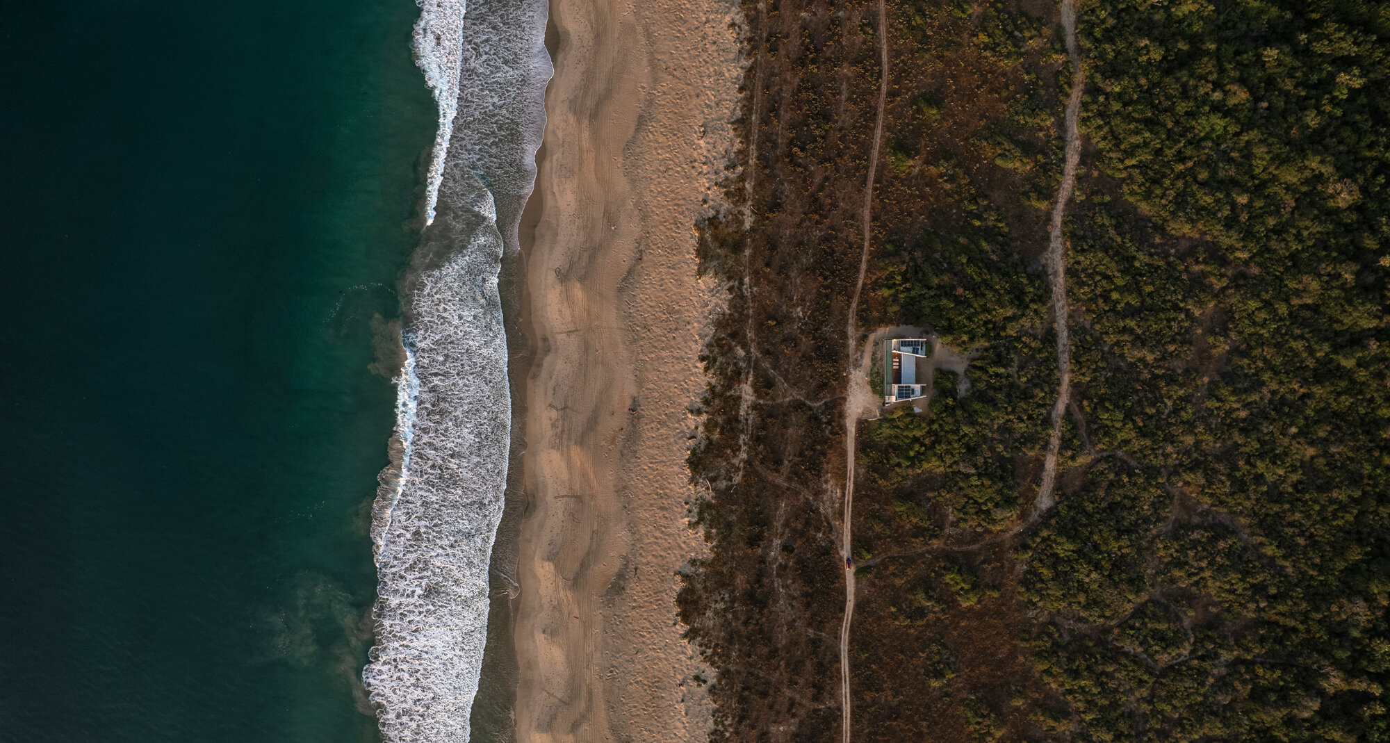 Aerial view of a small house in dense coastal scrub, set back from a sandy beach and breaking waves