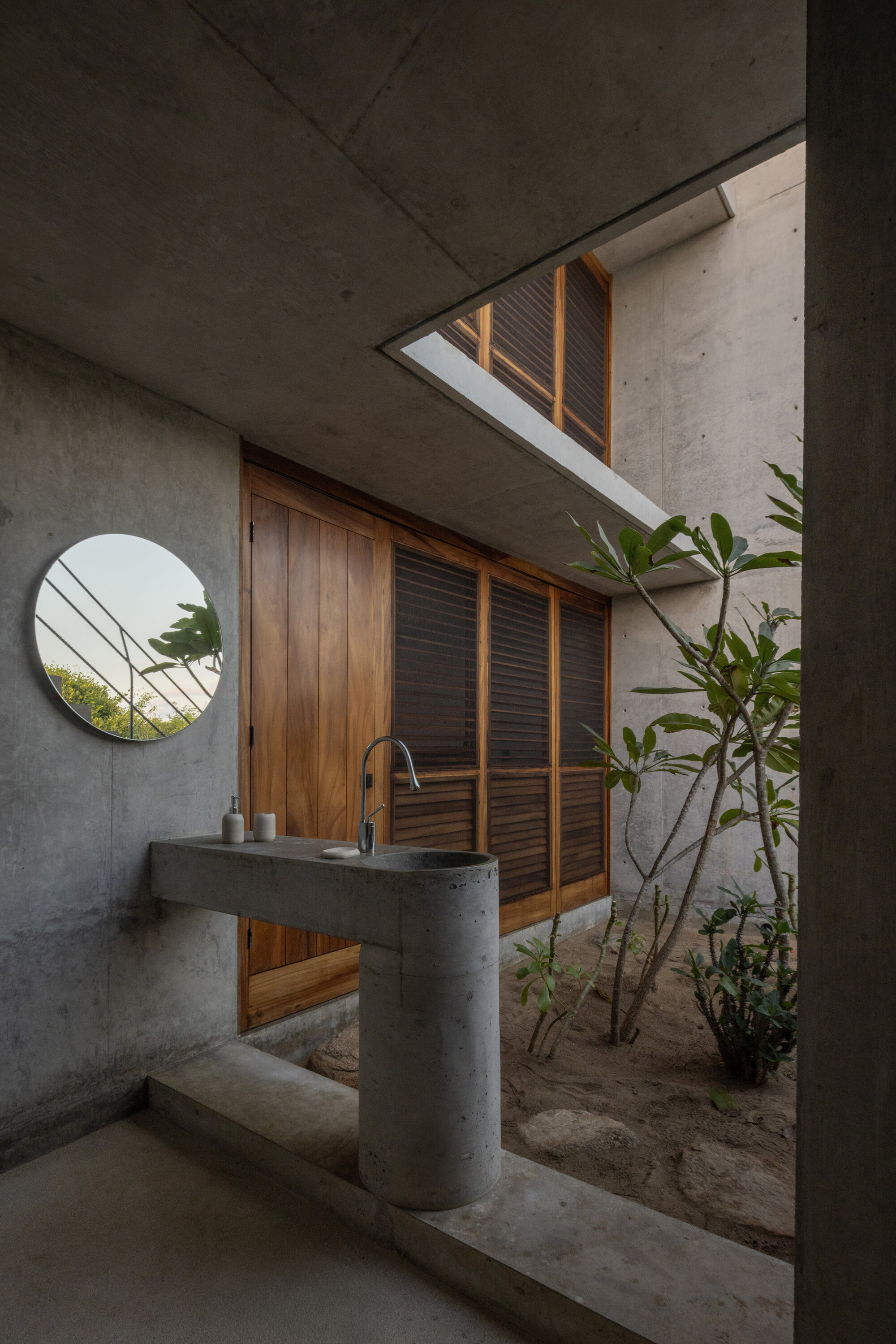 Round-ended concrete basin on a cylindrical pedestal beside a frangipani tree, with timber louvered doors and a circular mirror