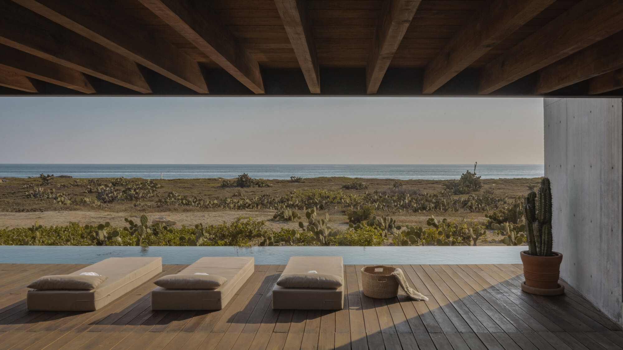 Timber deck with three cushioned loungers beneath exposed beams, looking across a pool to cactus scrub and the sea