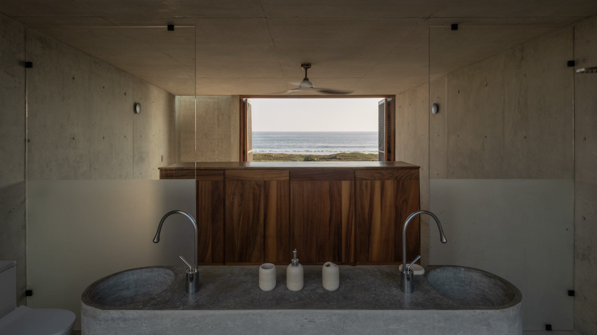 Concrete bathroom with a twin stone trough basin and walnut cabinet framing a window onto the sea