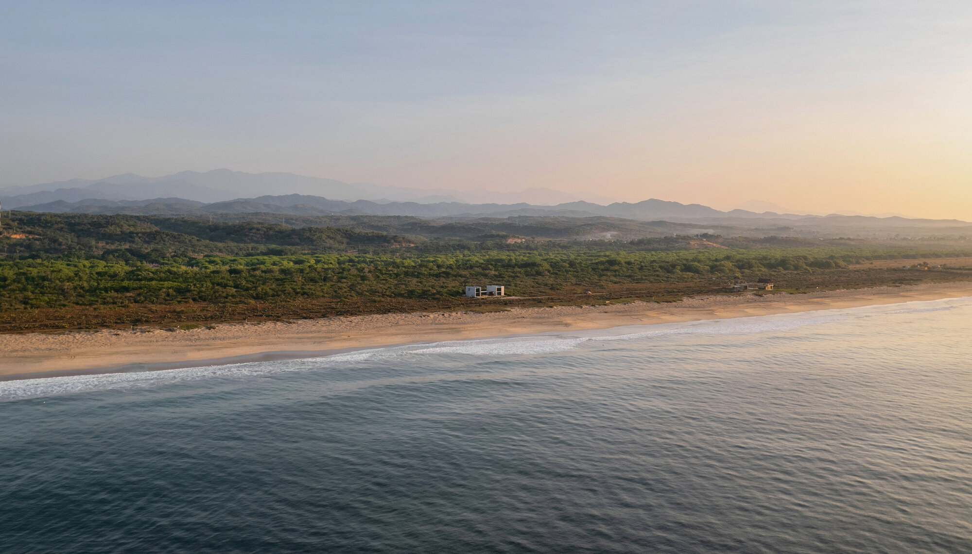 Aerial view of a small concrete house on a long sandy beach backed by forested hills in morning haze