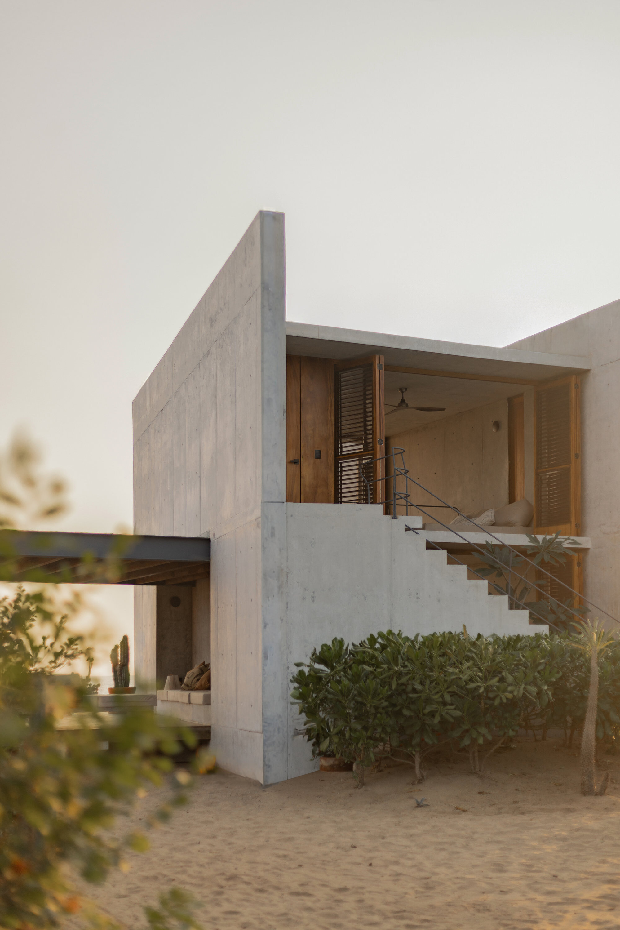 Board-formed concrete house with an exterior stair rising to a shuttered timber upper room, seen across sand in warm evening haze