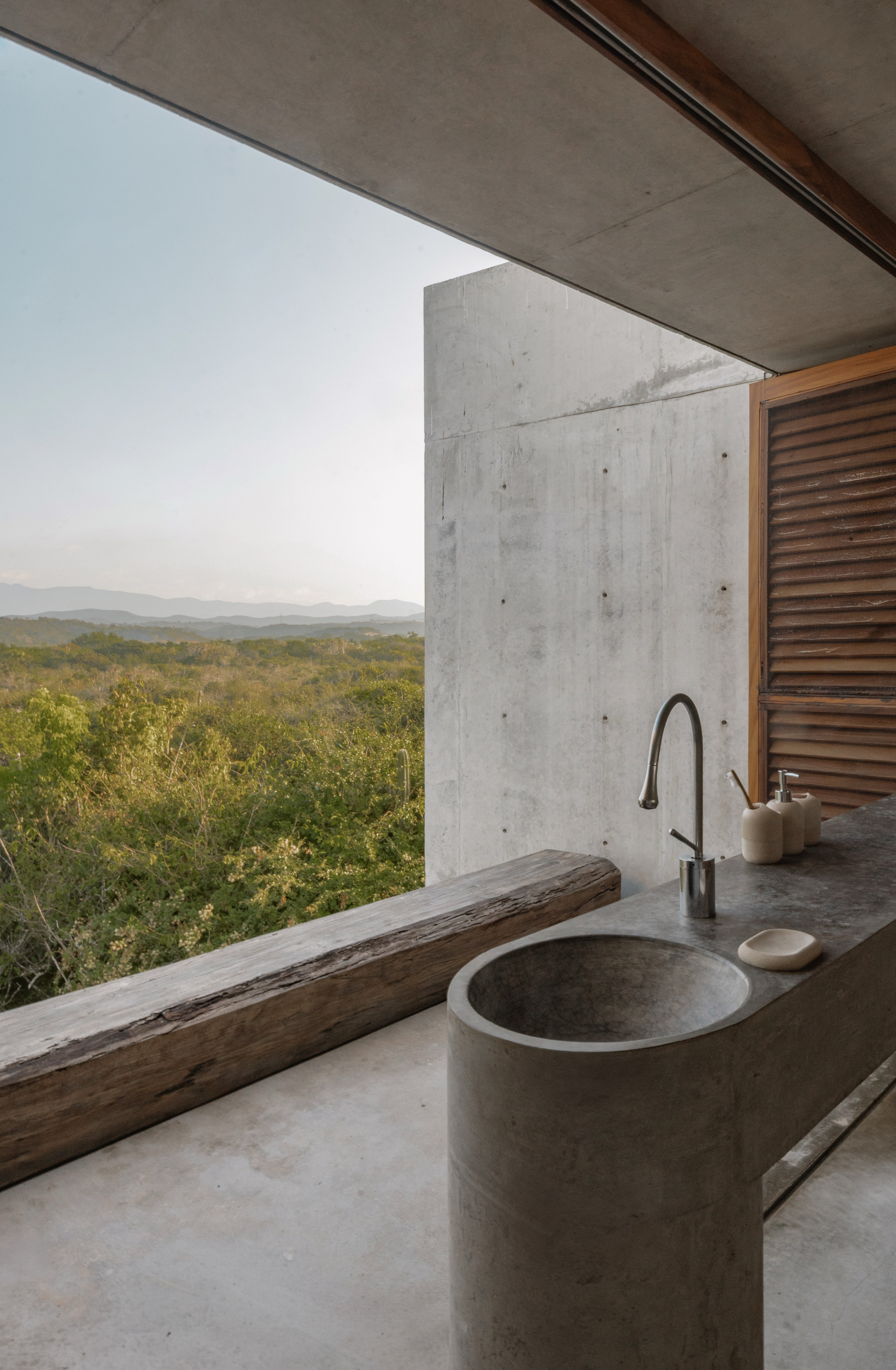 Round concrete basin and counter beside a reclaimed timber ledge, opening onto green scrubland and distant hills at dusk
