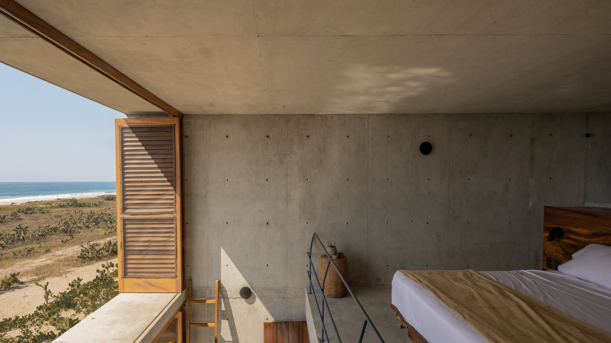 Concrete bedroom with a folded-back timber louvre shutter and a bed facing the ocean and dune vegetation in bright daylight
