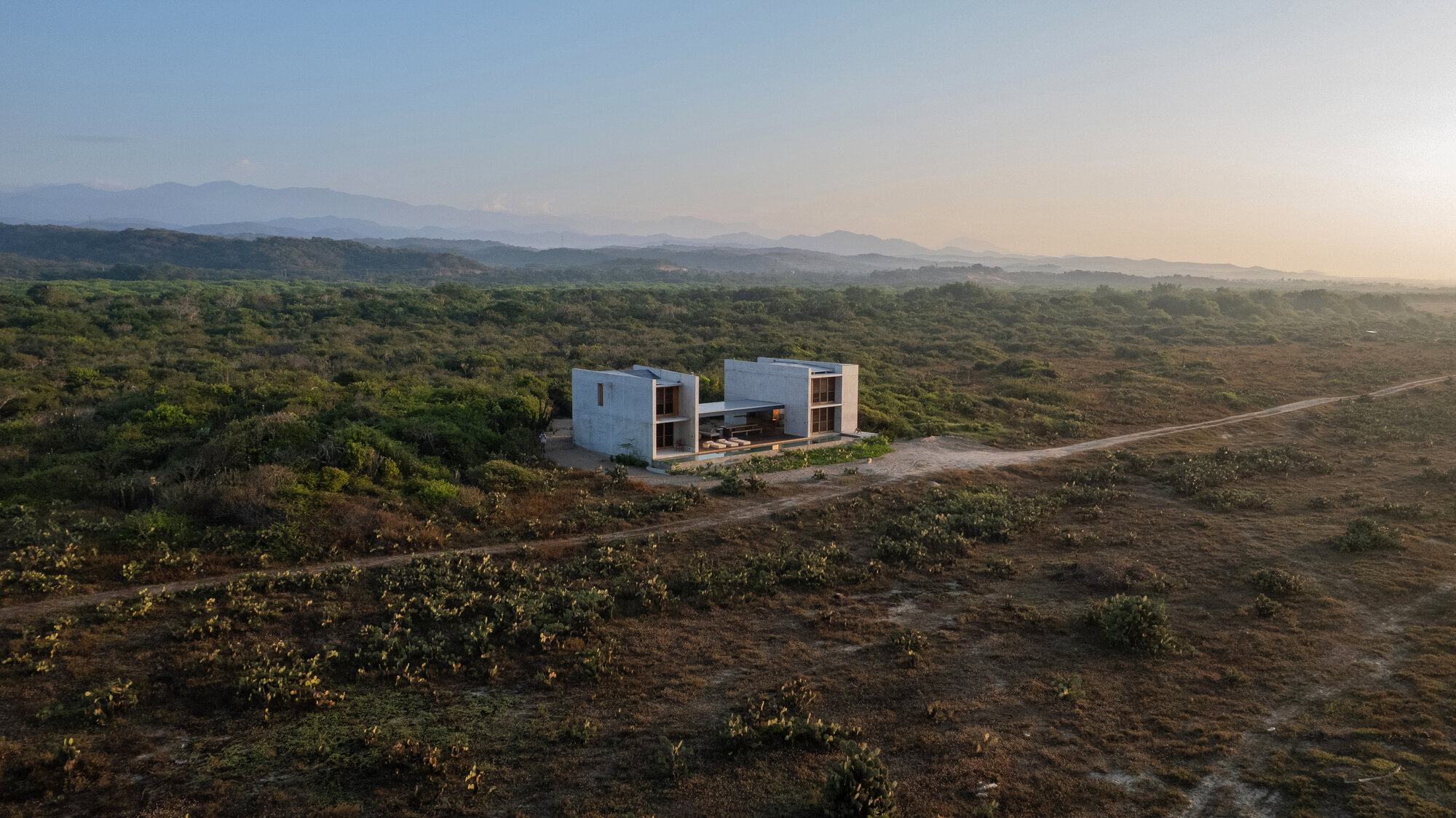 Drone view of two concrete blocks joined by a covered porch, set in dense scrub with a dirt track and hazy mountains