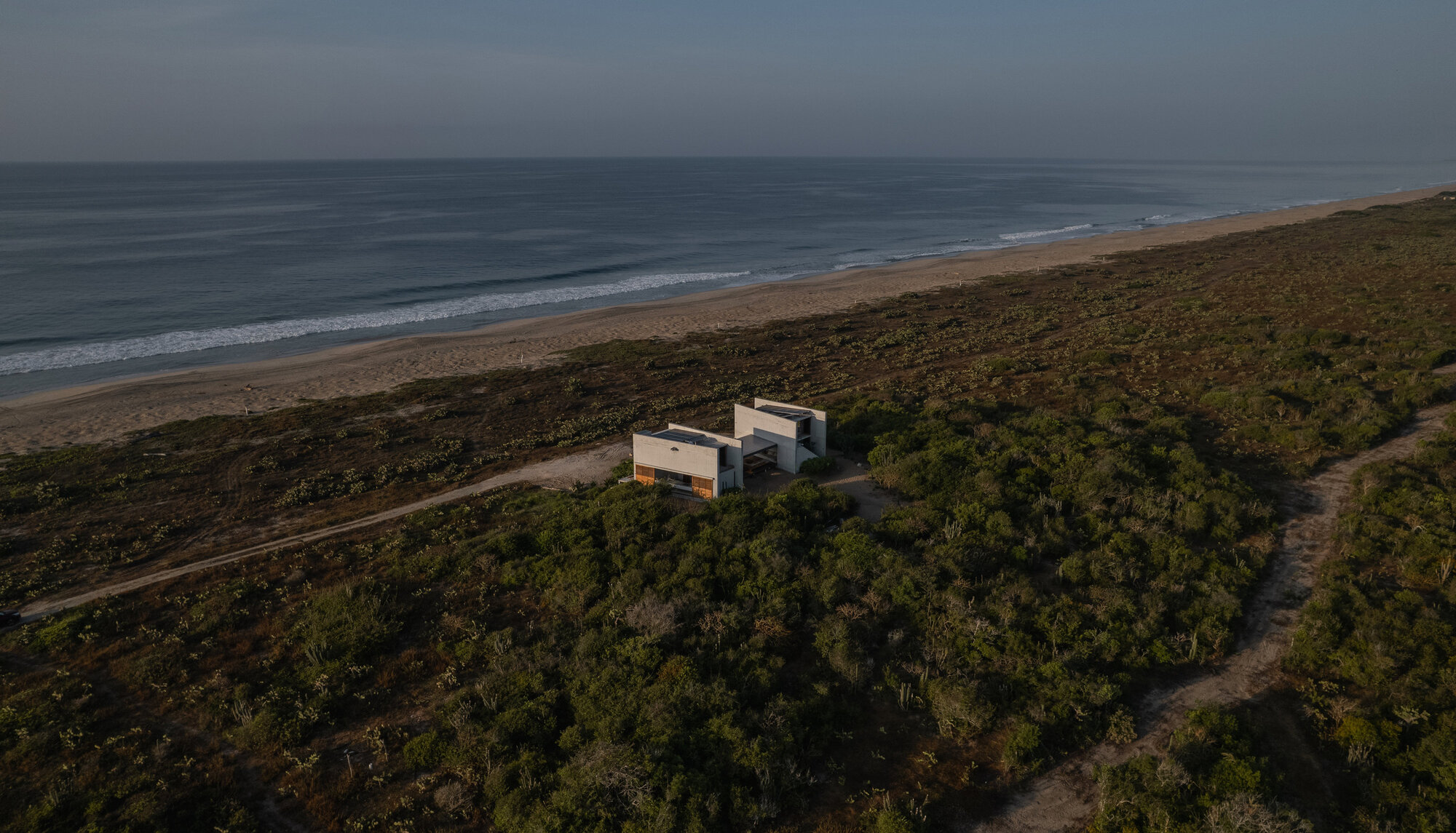 Aerial view of the white concrete house set among coastal scrub, with a sandy track and surf along the beach at dusk