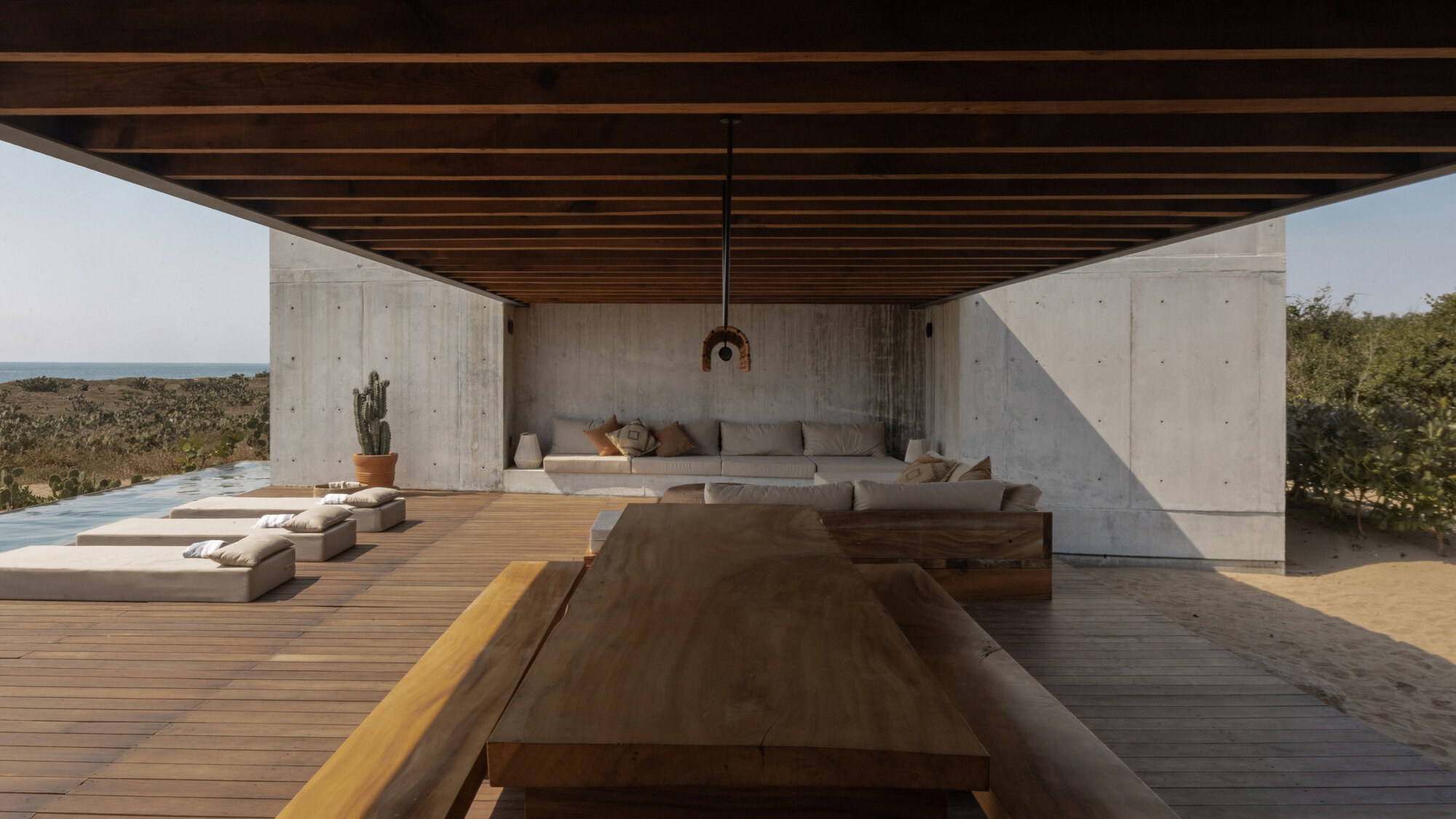 Open concrete pavilion with timber ceiling beams, built-in sofa, plank dining table and daybeds beside a lap pool in hazy sun