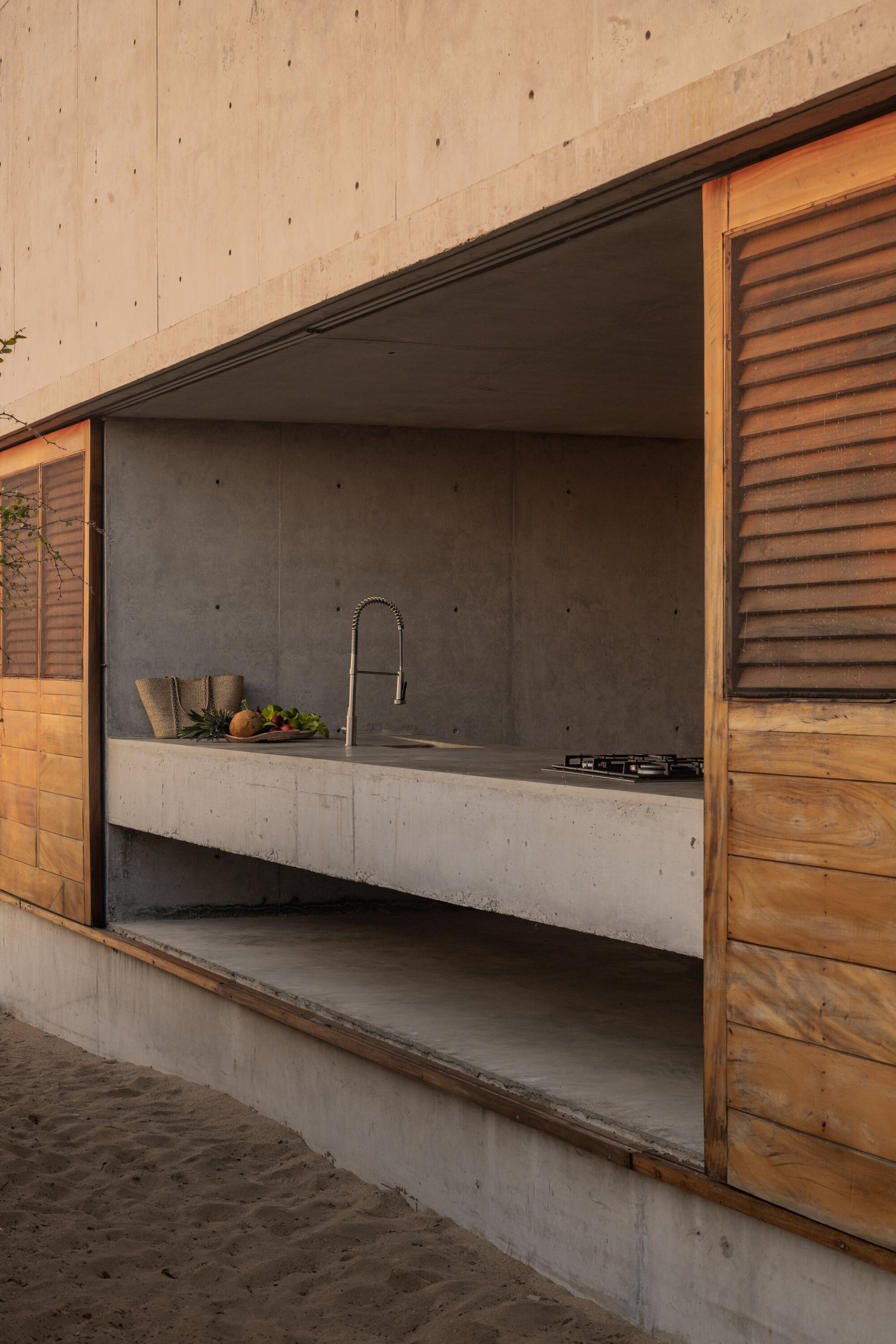 Outdoor kitchen opening in a concrete wall with a sliding louvered timber shutter, steel faucet and cooktop, warm light on the sand