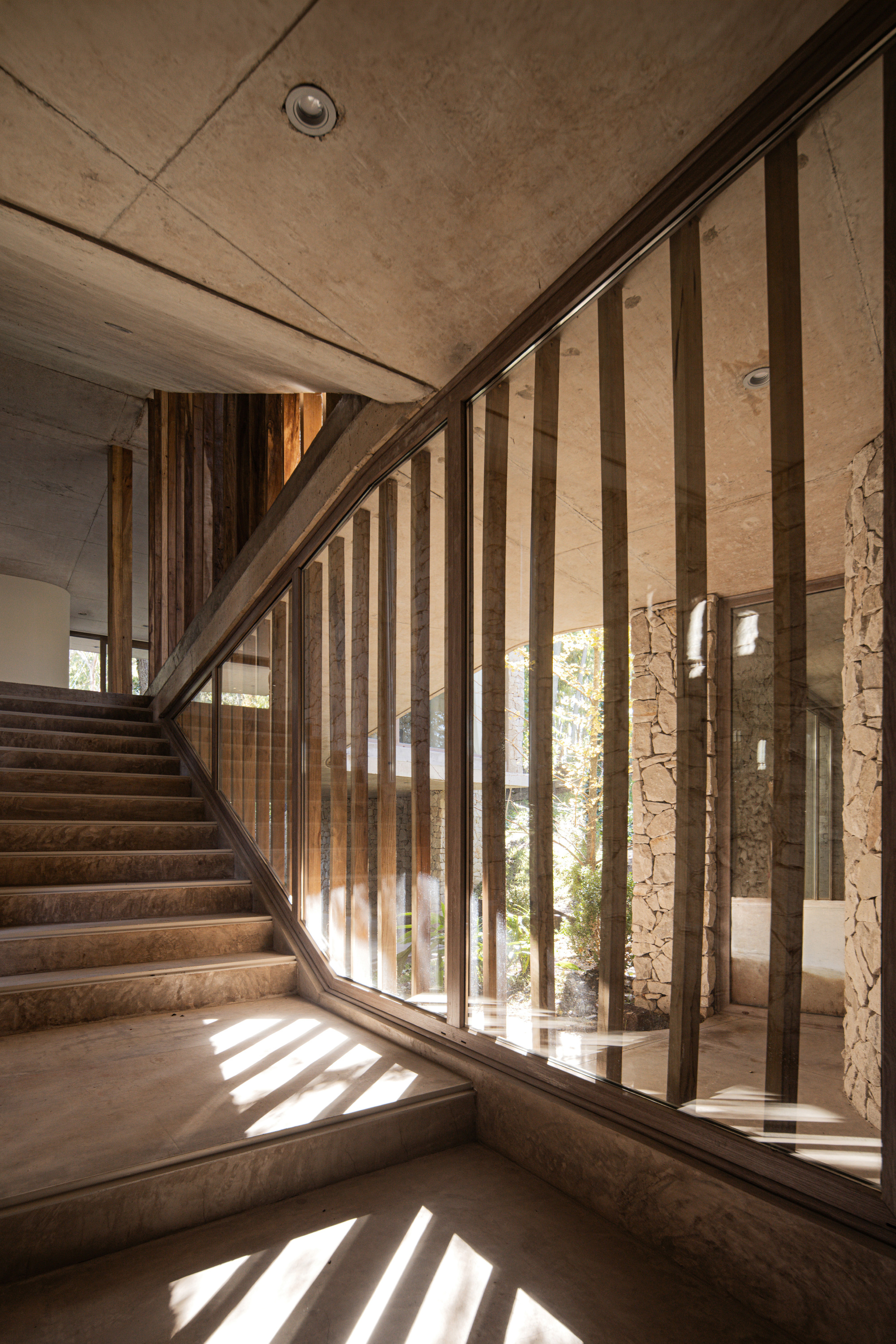 Concrete stair beside a glass wall with vertical timber fins, throwing striped sunlight across the landing and stone walls beyond