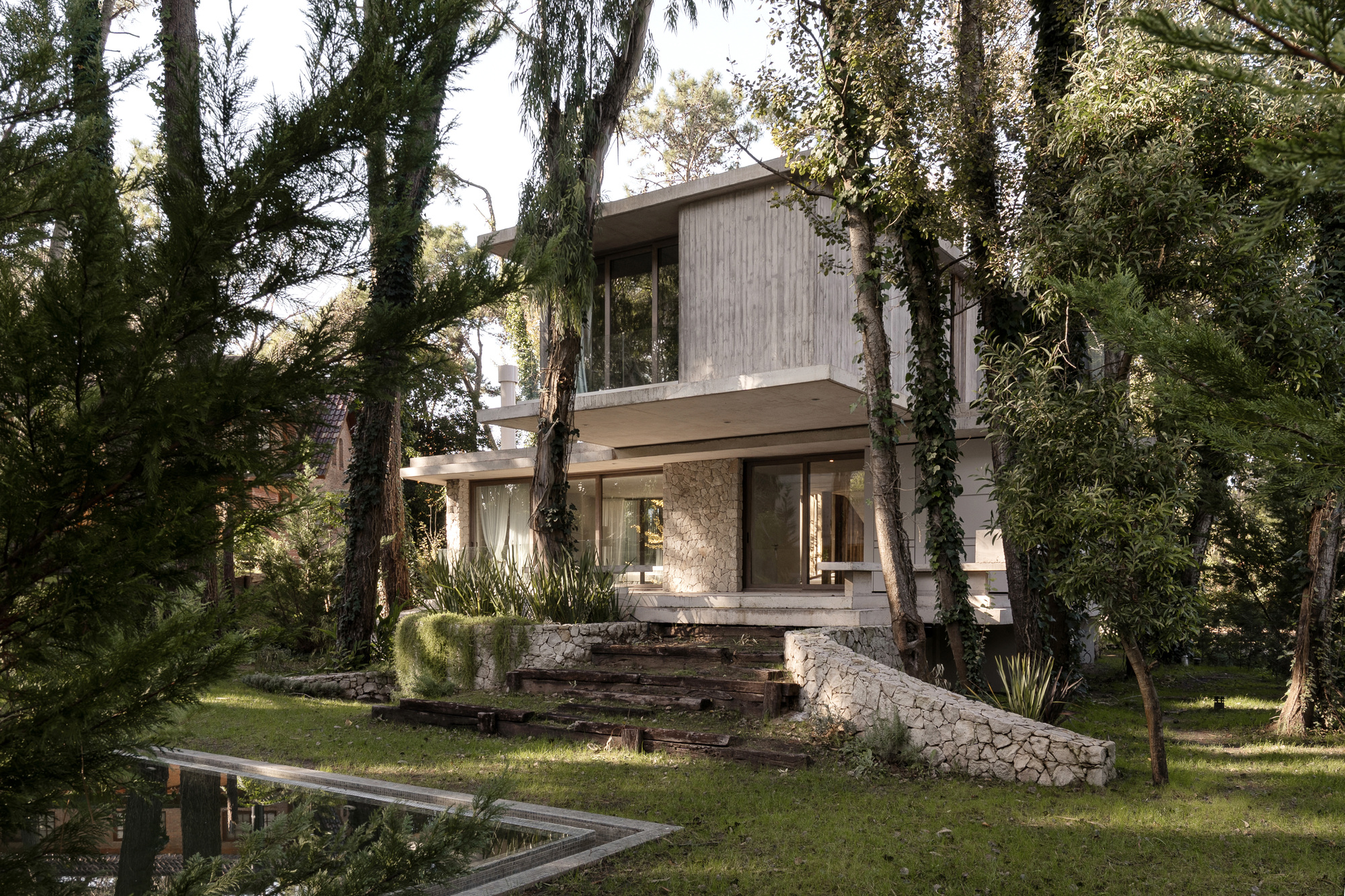Two-storey concrete house behind a stone retaining wall and timber steps, set among tall trees on a lawn