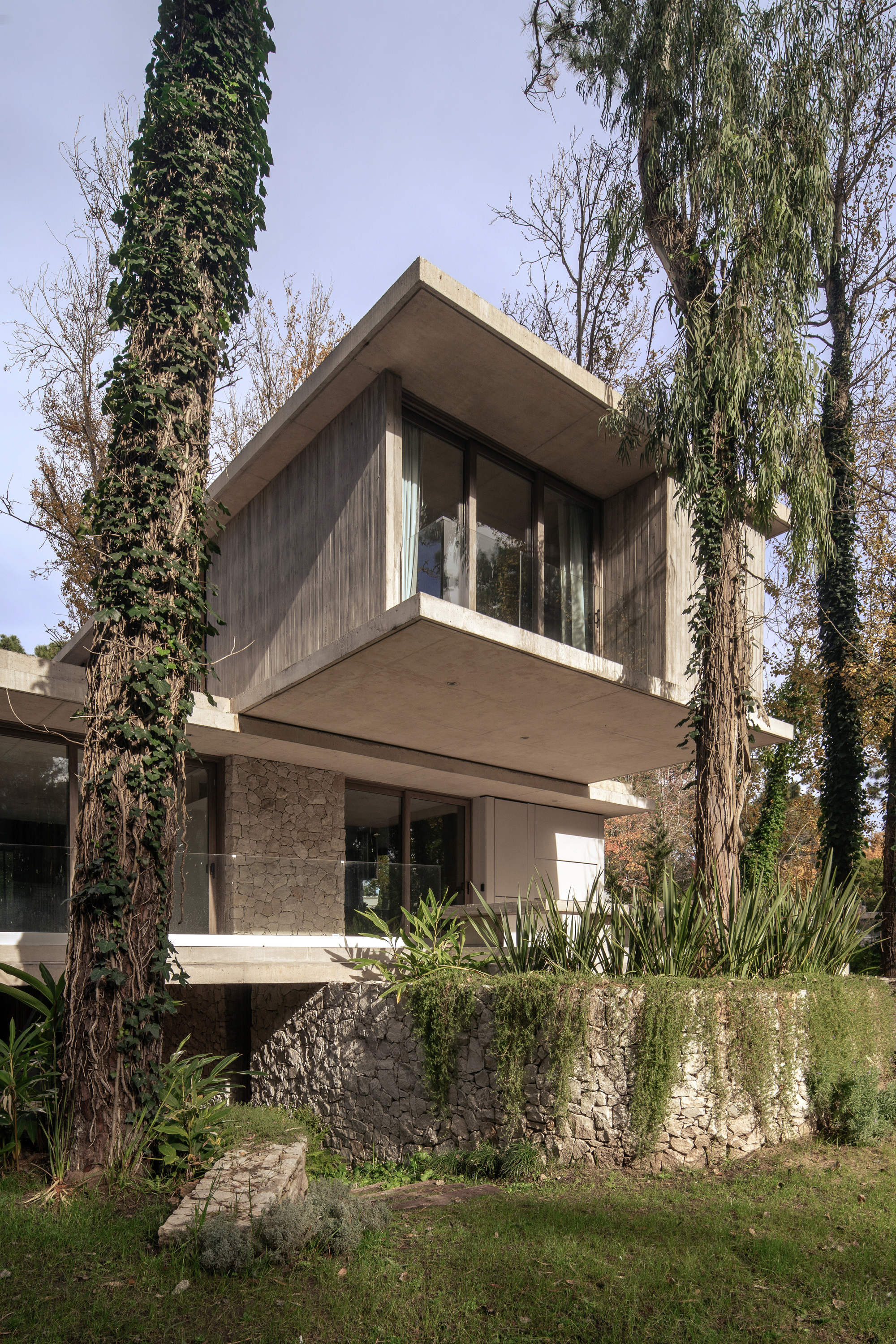 Stacked concrete volumes cantilevering over a rubble stone base, flanked by ivy-covered tree trunks under a cloudy sky