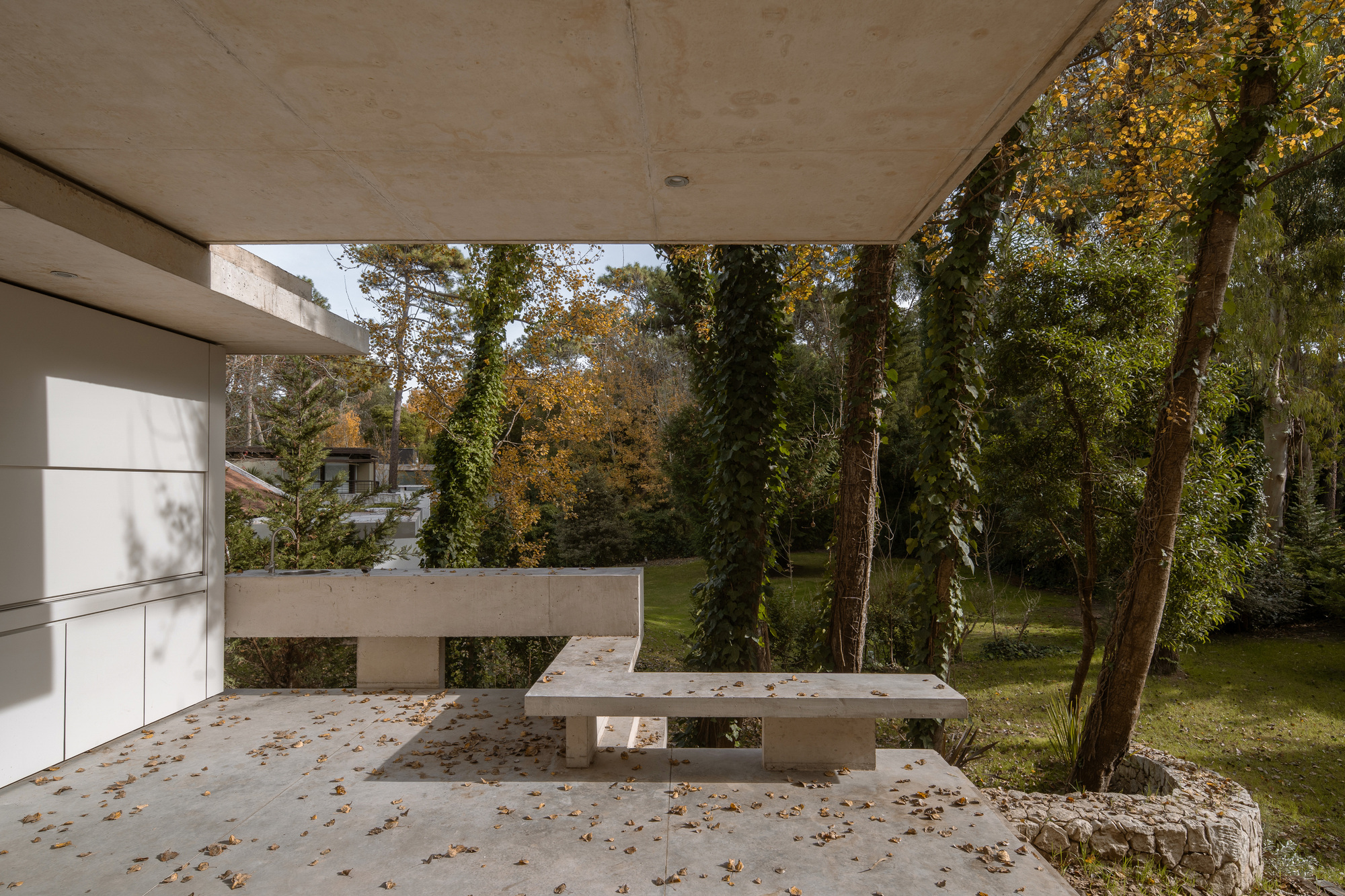 Covered concrete terrace with a built-in bench and low wall, scattered with fallen leaves among ivy-wrapped trees in autumn