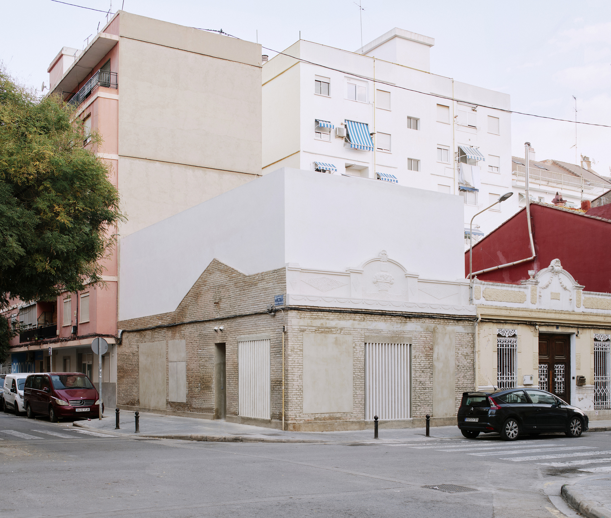 Street corner view of a brick building topped by a white rendered volume, with parked cars and a tree in soft daylight