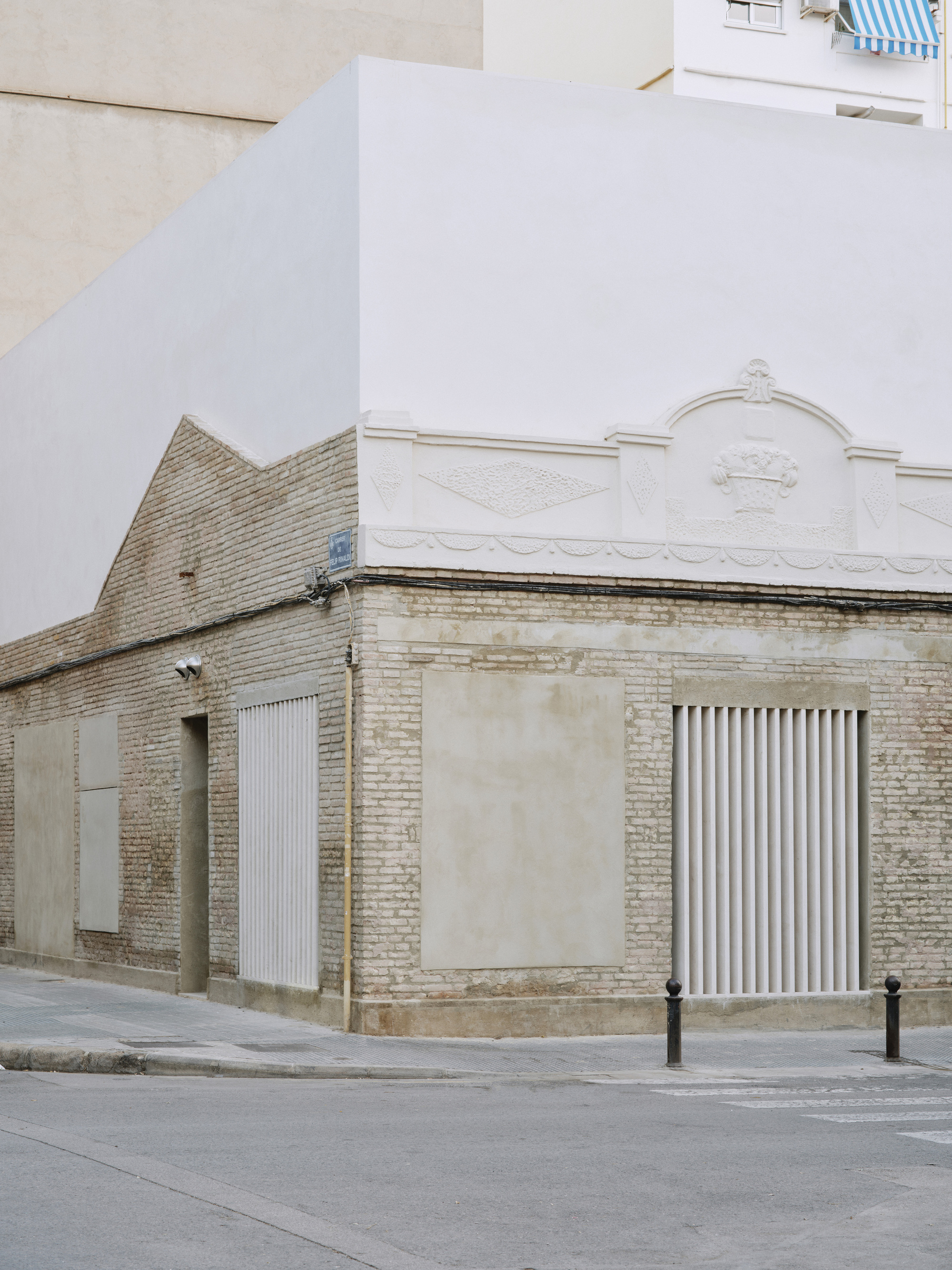 Corner of the old brick facade with plaster pediment, shuttered openings and a white box above, seen from an empty street