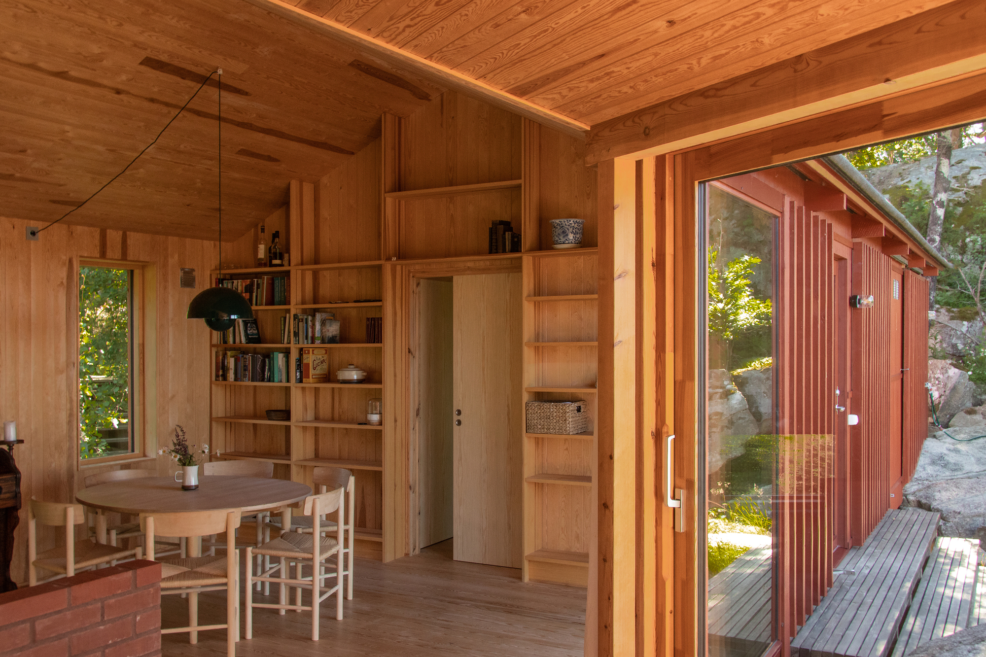 Timber-lined dining room with built-in bookshelves, round table and a sliding glass door opening onto a red-clad wall and rock outcrop