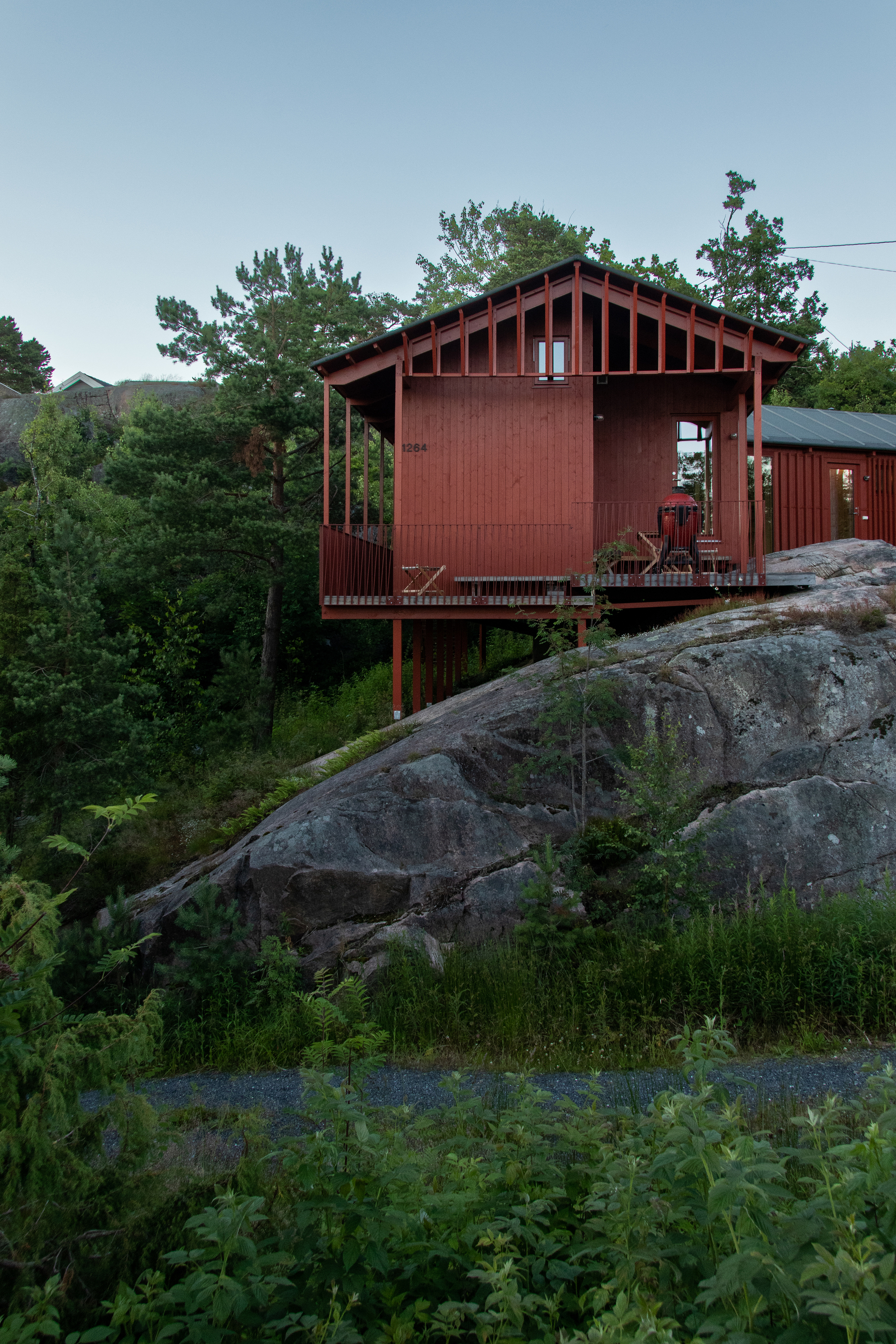 Red timber cabin raised on stilts above a granite outcrop, framed by pines in soft evening light