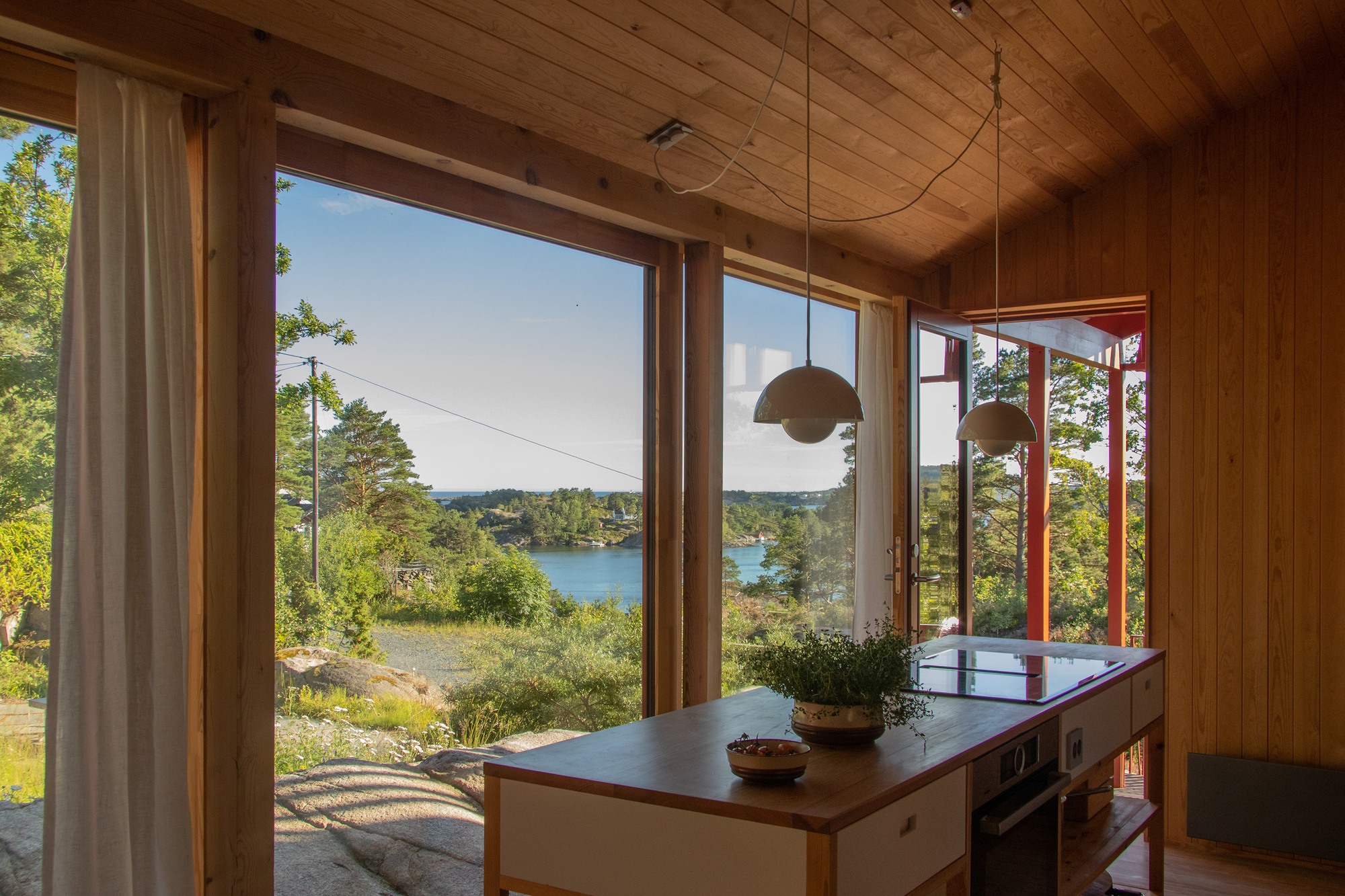 Kitchen island with induction hob and potted herbs before tall windows overlooking a sunlit rocky inlet