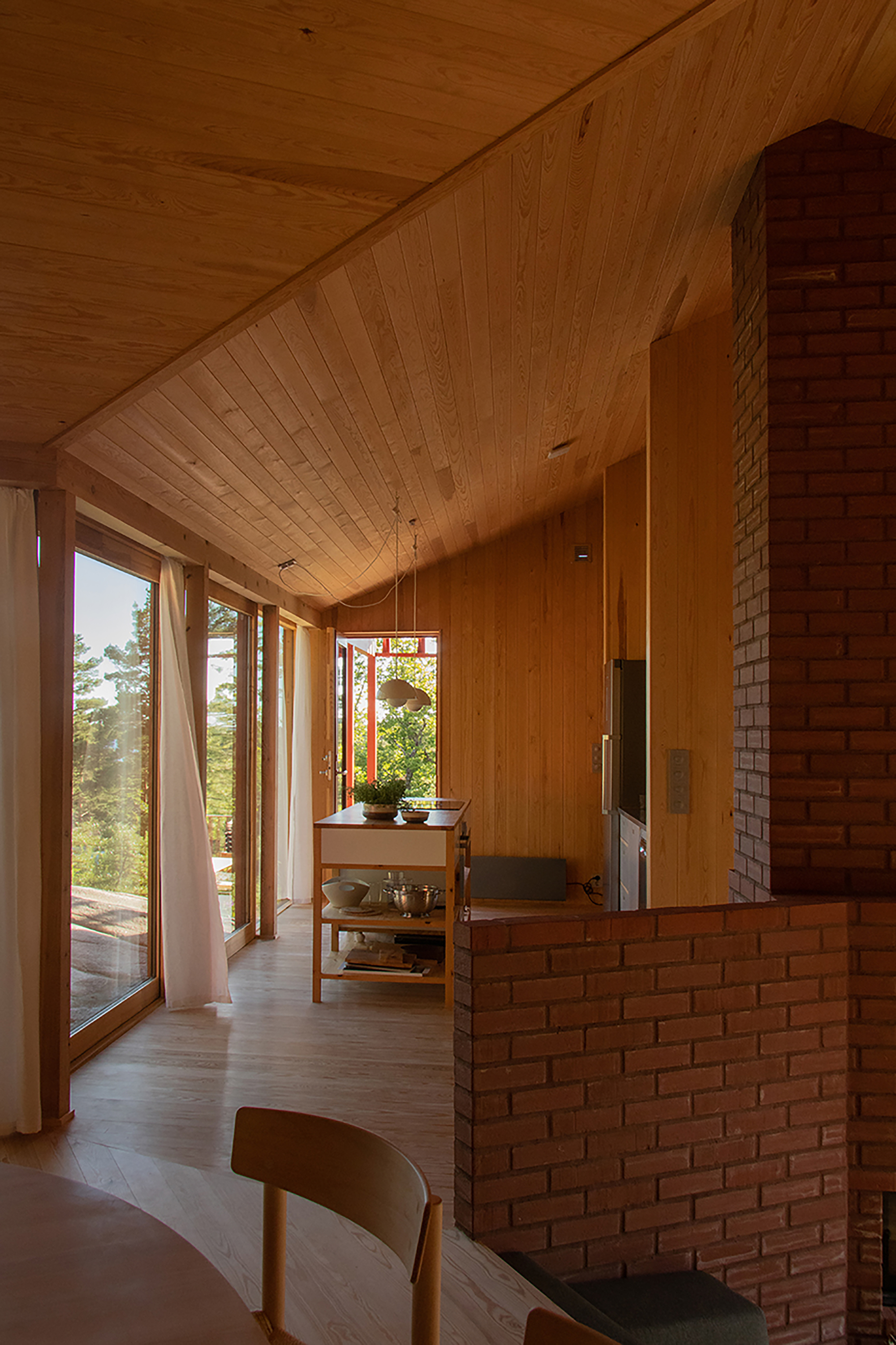 Pine-lined interior with sloped ceiling, brick chimney mass and floor-to-ceiling windows with a billowing white curtain