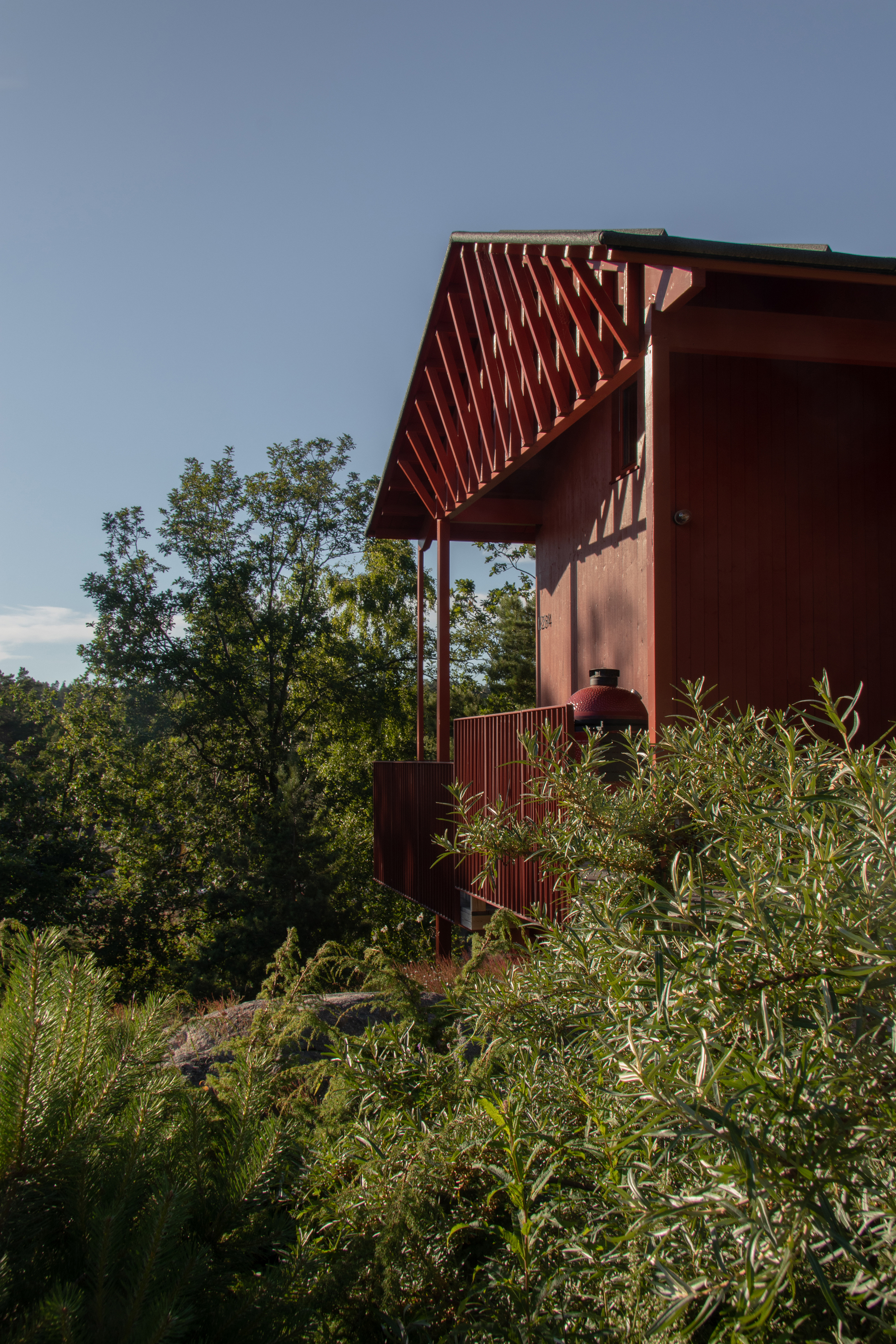Red-painted cabin corner with exposed rafters and slim posts, partly hidden behind shrubs under a clear blue sky