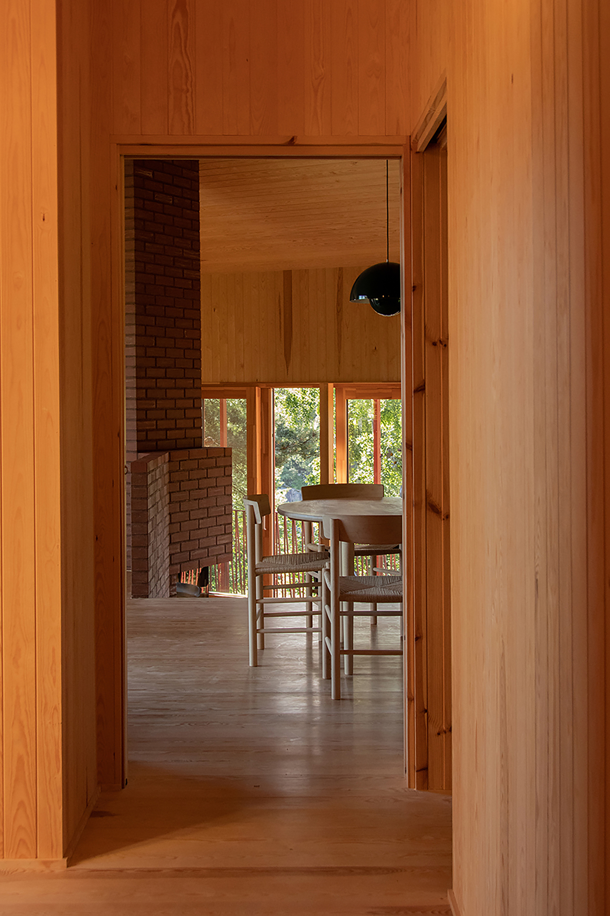 Doorway view into a timber-lined dining room with a brick fireplace, round table and woven chairs, lit by green forest light