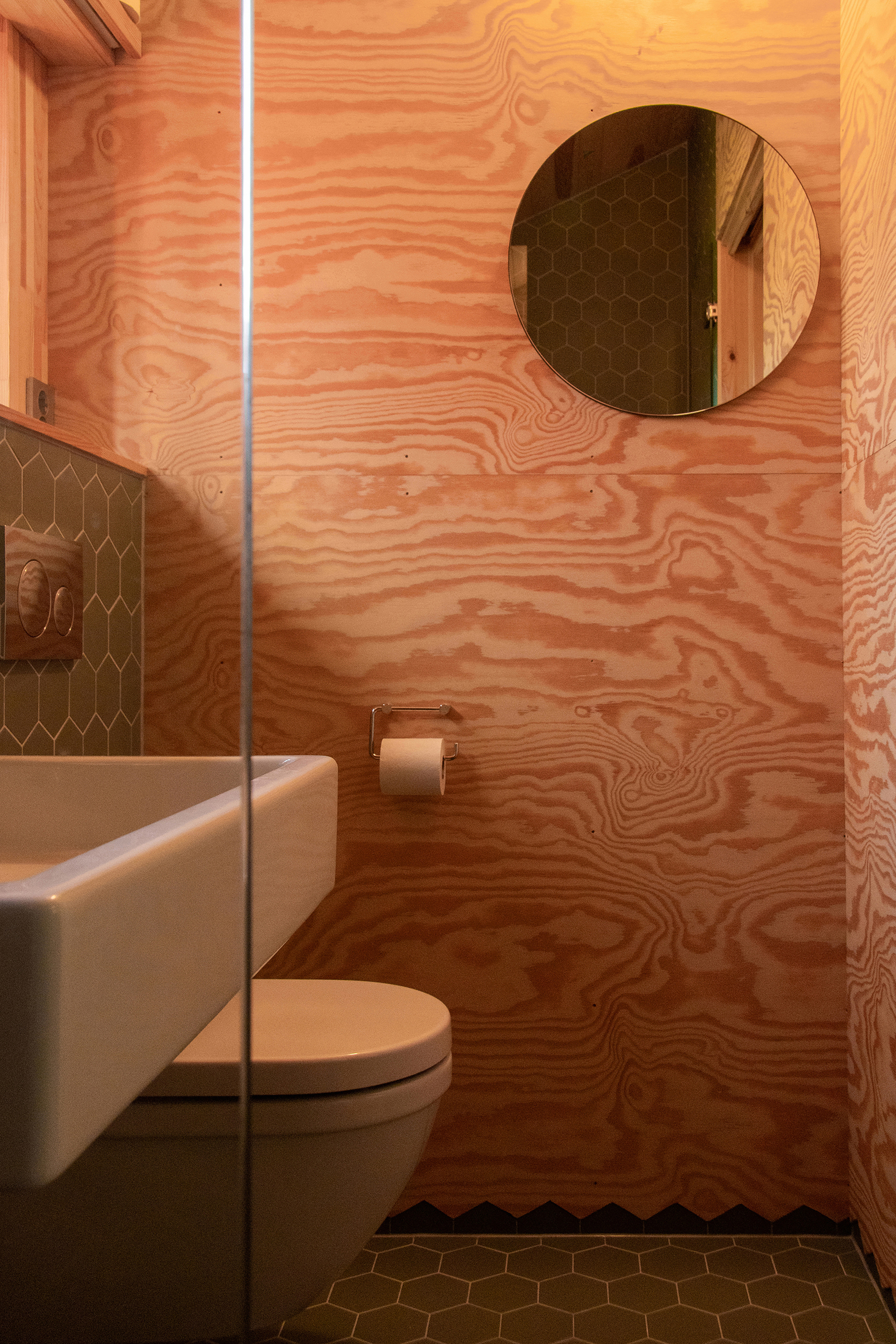 Small bathroom with figured plywood walls, a round mirror and green hexagonal tiles under warm lamplight