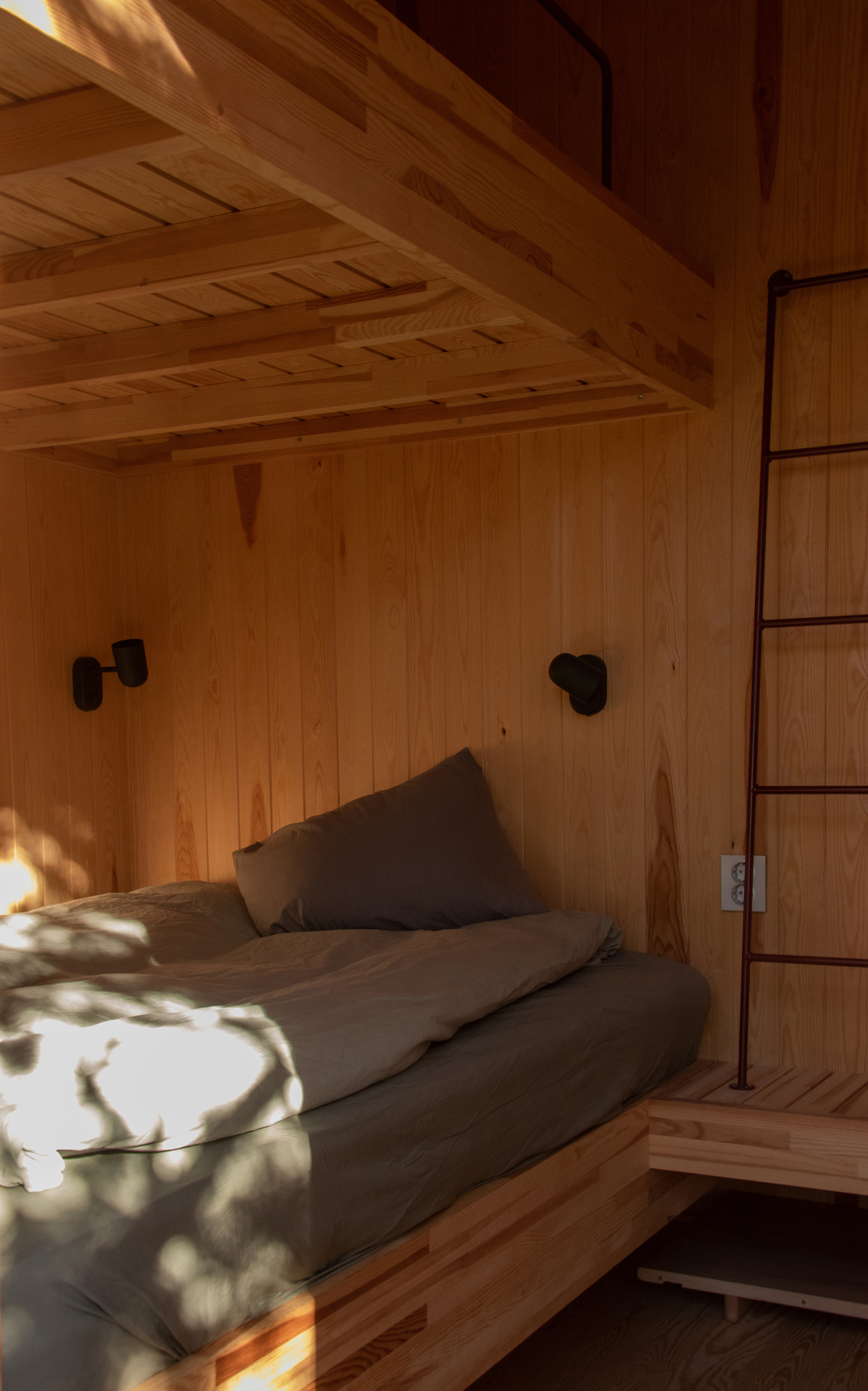 Built-in timber bunk with grey bedding, black wall lamps and a steel ladder, dappled with afternoon sunlight through leaves