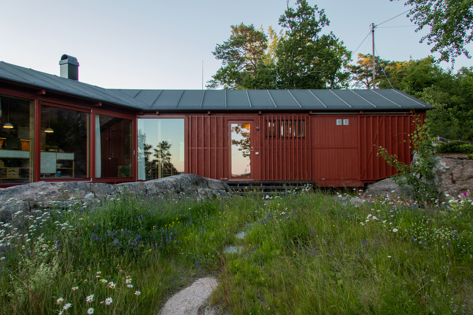 Red timber cabin with glazed corner and metal roof behind a wildflower meadow and granite boulders at dusk